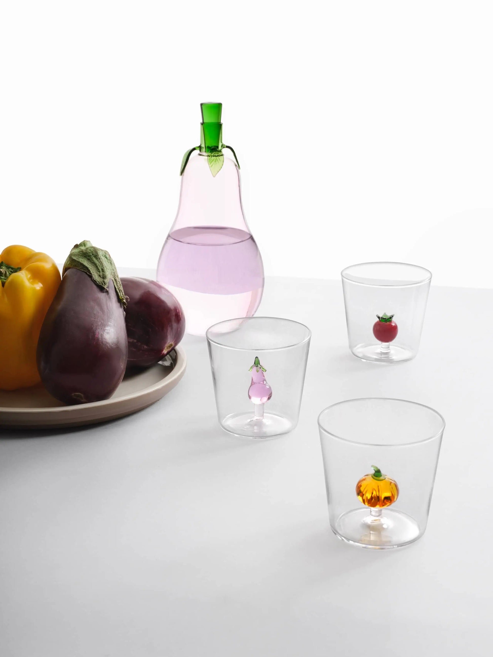 A handcrafted plate with yellow peppers and eggplants sits beside three clear glasses, each holding a small fruit or vegetable figurine, and an Ichendorf Milano Vegetables Bottle filled with pink liquid and topped with green.
