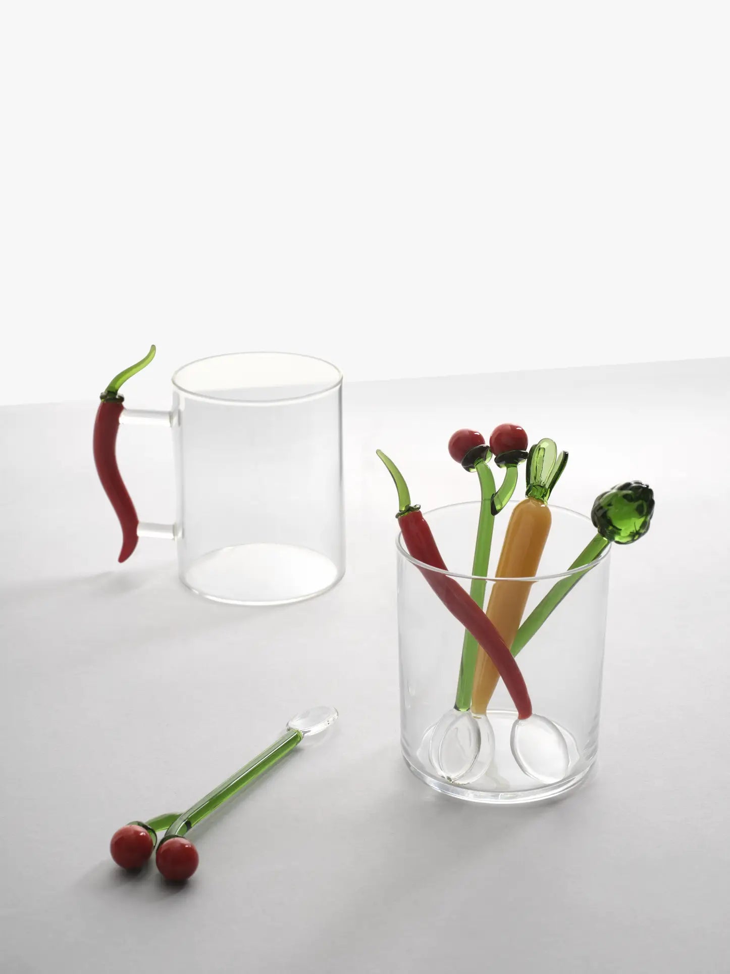 Two clear glass cups: one features a red chili pepper-shaped handle, the other holds Ichendorf Milano’s Vegetables Spoons Set of 2. A glass cherry stirring stick by Ichendorf Milano rests on the table nearby.