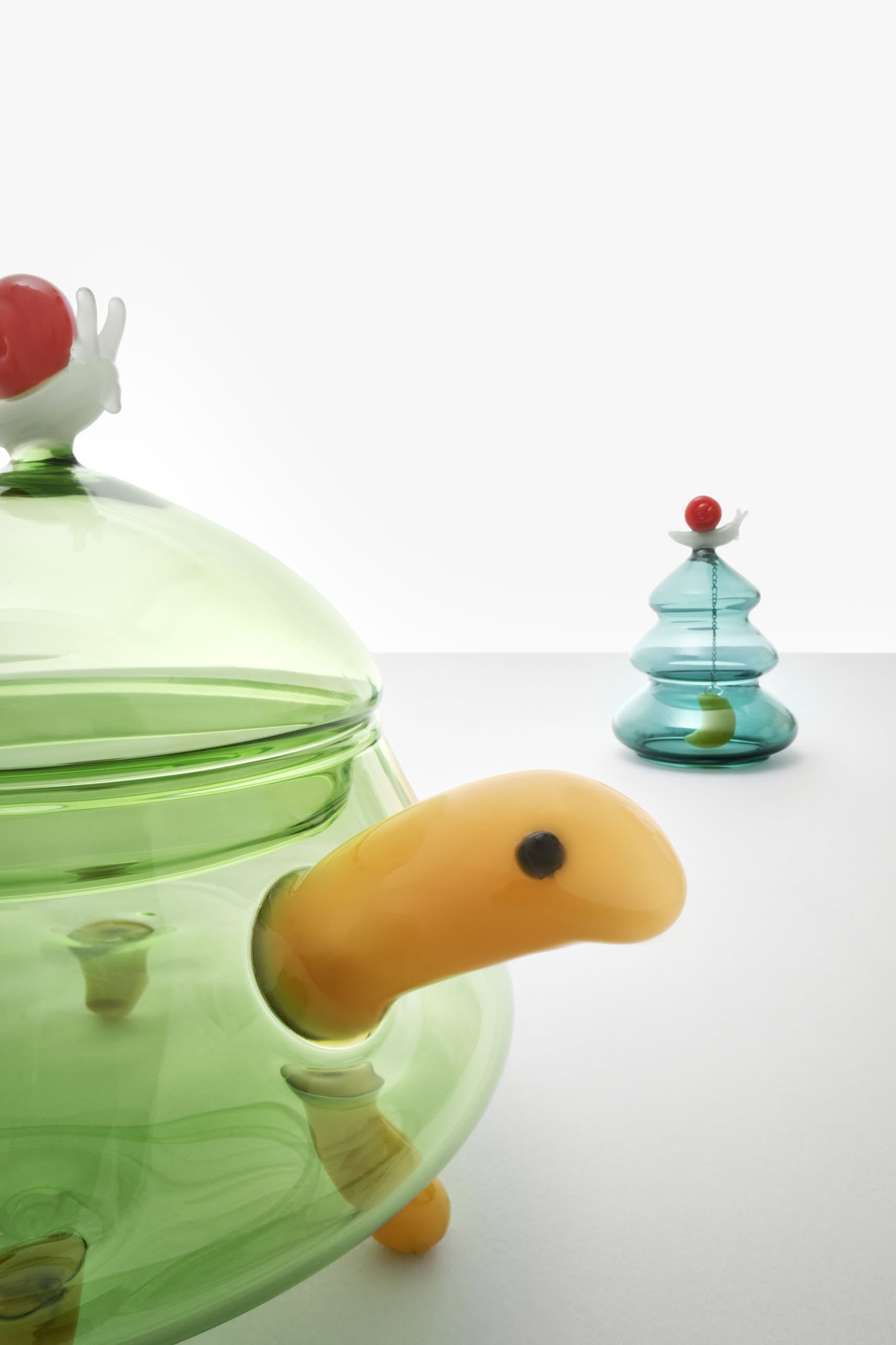 The Ichendorf Milano Woodland Tales Sugar Bowl by Ichendorf Milano features a green glass turtle with a yellow head and red lid detail, set against a blue tree-shaped glass object for a magical, nature-inspired touch.