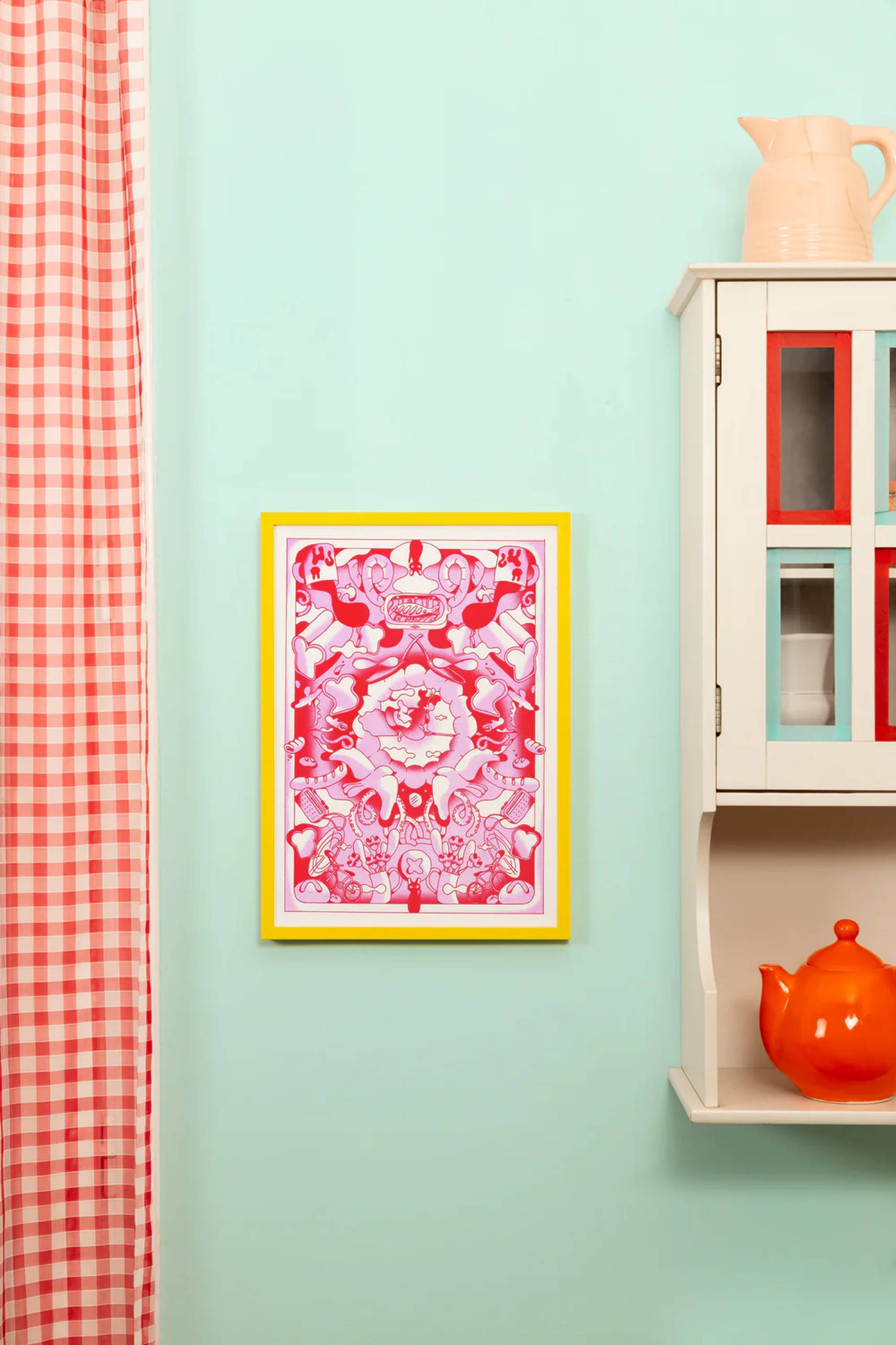 The KIBLIND Atelier Kiki La Petite Sorciere art print by Agathe Bruguiere hangs in a yellow frame on a mint green wall beside a red and white checked curtain and a white kitchen cabinet with colorful dishes and an orange teapot.