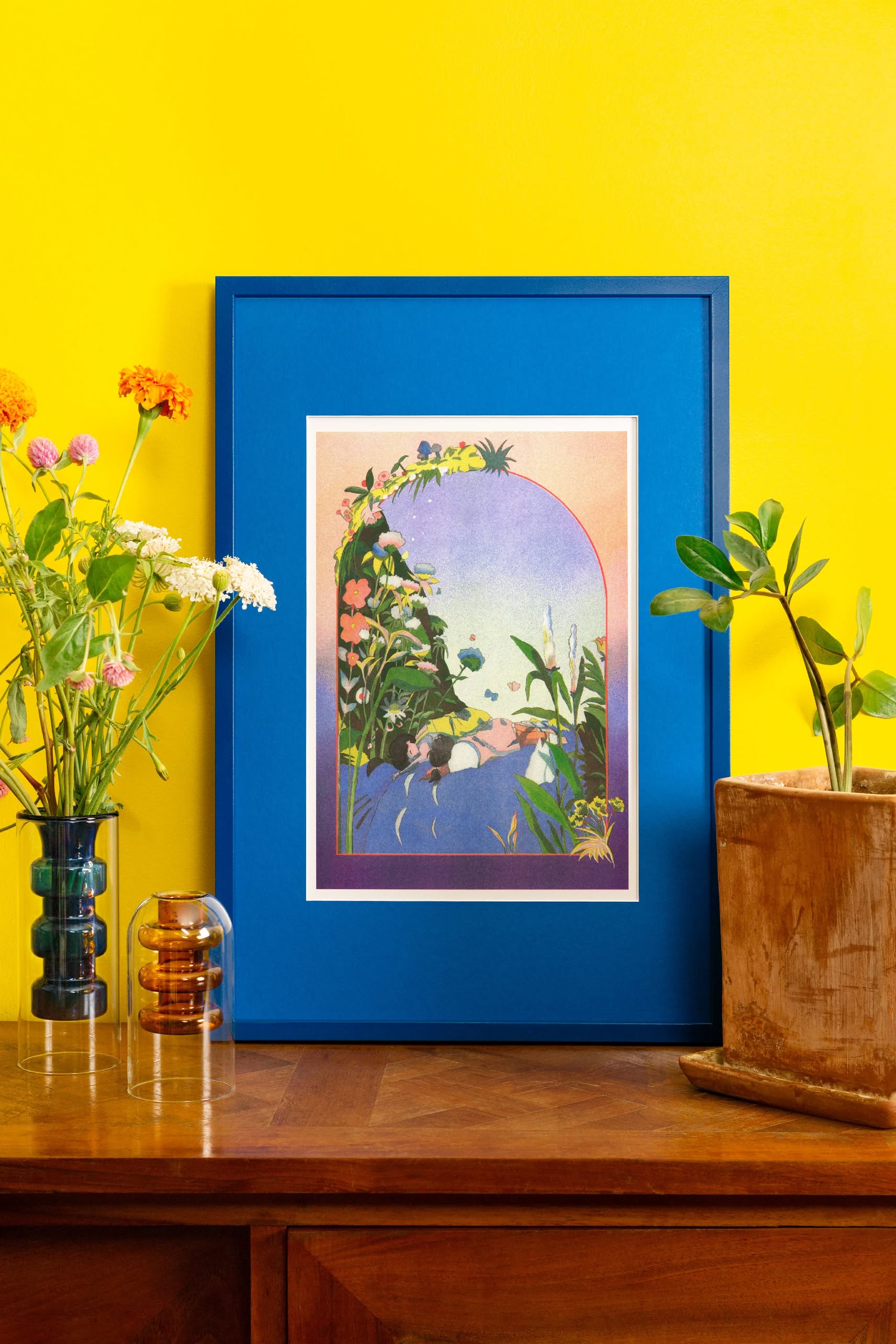The KIBLIND Atelier Jeanne Saboureault - My Neighbor Totoro risoprint sits on a wooden table against a yellow wall, its bright blue frame surrounded by vases of flowers and a potted plant.