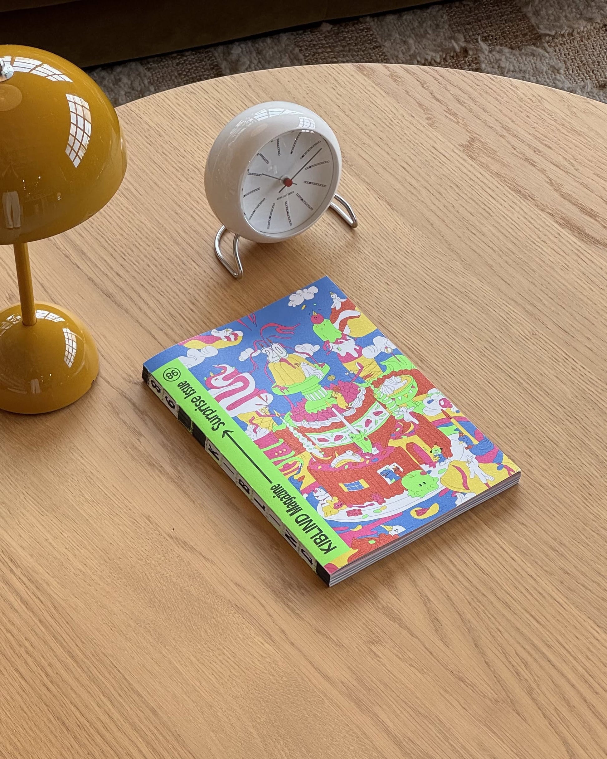 KIBLIND Atelier Magazine #86 by KIBLIND Atelier, with its vivid cover, rests on a light wood table beside a small white clock and yellow lamp, while a brown couch and part of a rug appear in the background.
