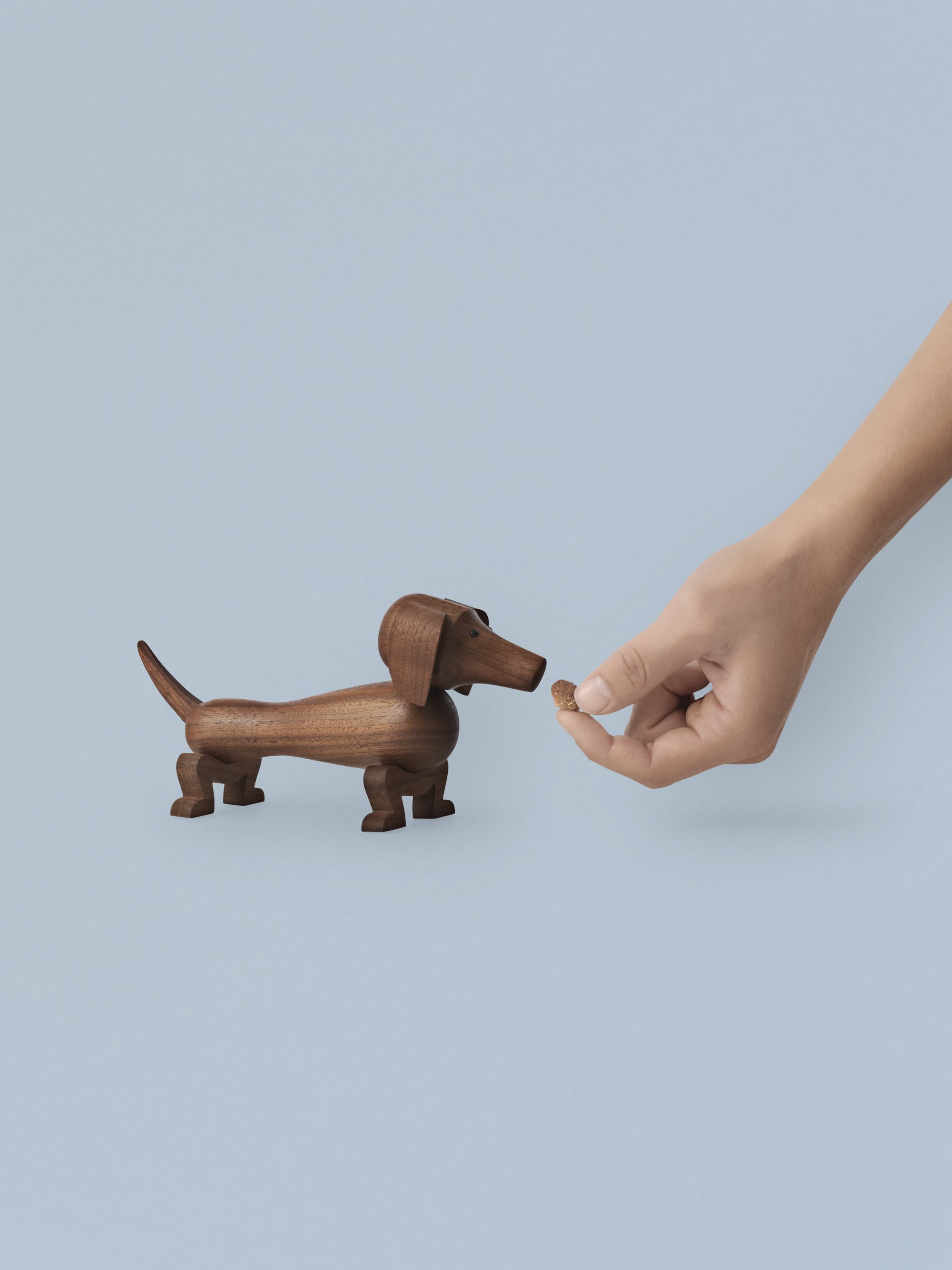 A hand holds a treat in front of a Kay Bojesen Dog Small by Kay Bojesen, the iconic wooden dog, set against a plain light blue background.