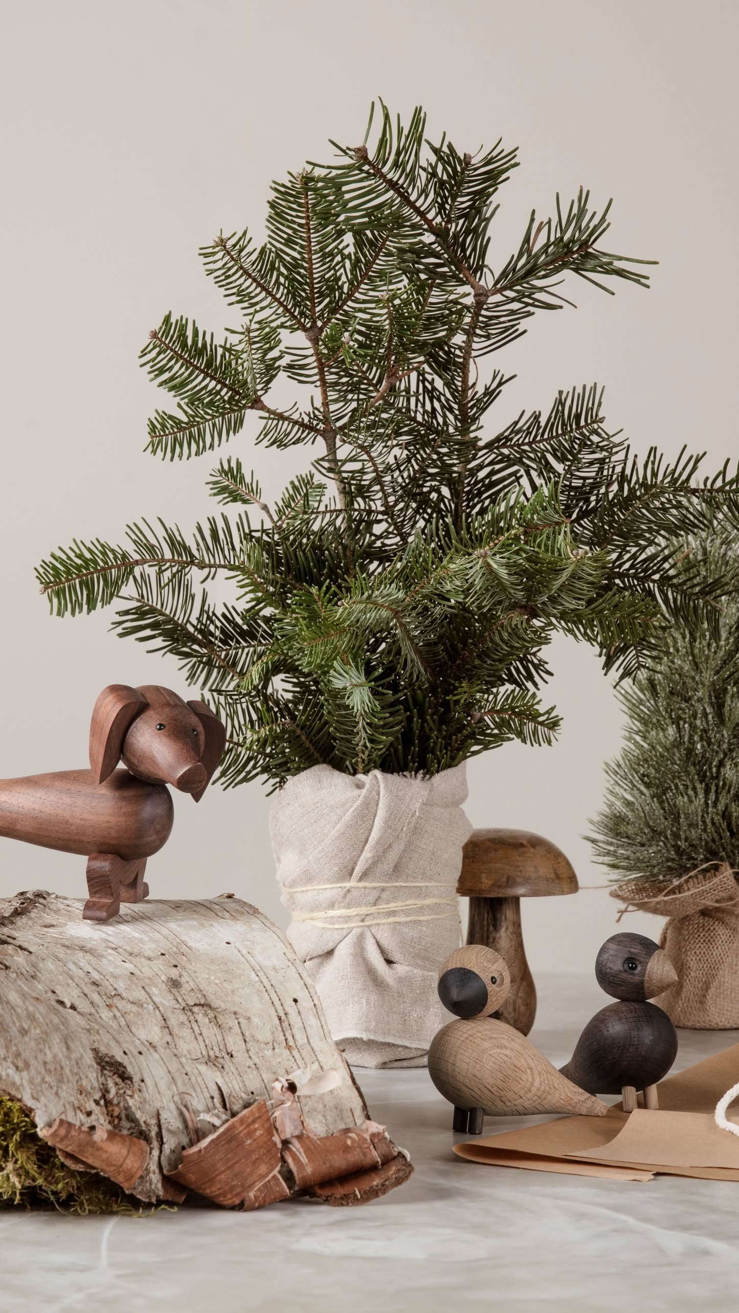 A small pine tree wrapped in fabric sits on a table with the Kay Bojesen Dog Small by Kay Bojesen, bird and mushroom figurines, some moss, and bark for a natural, rustic decor arrangement.