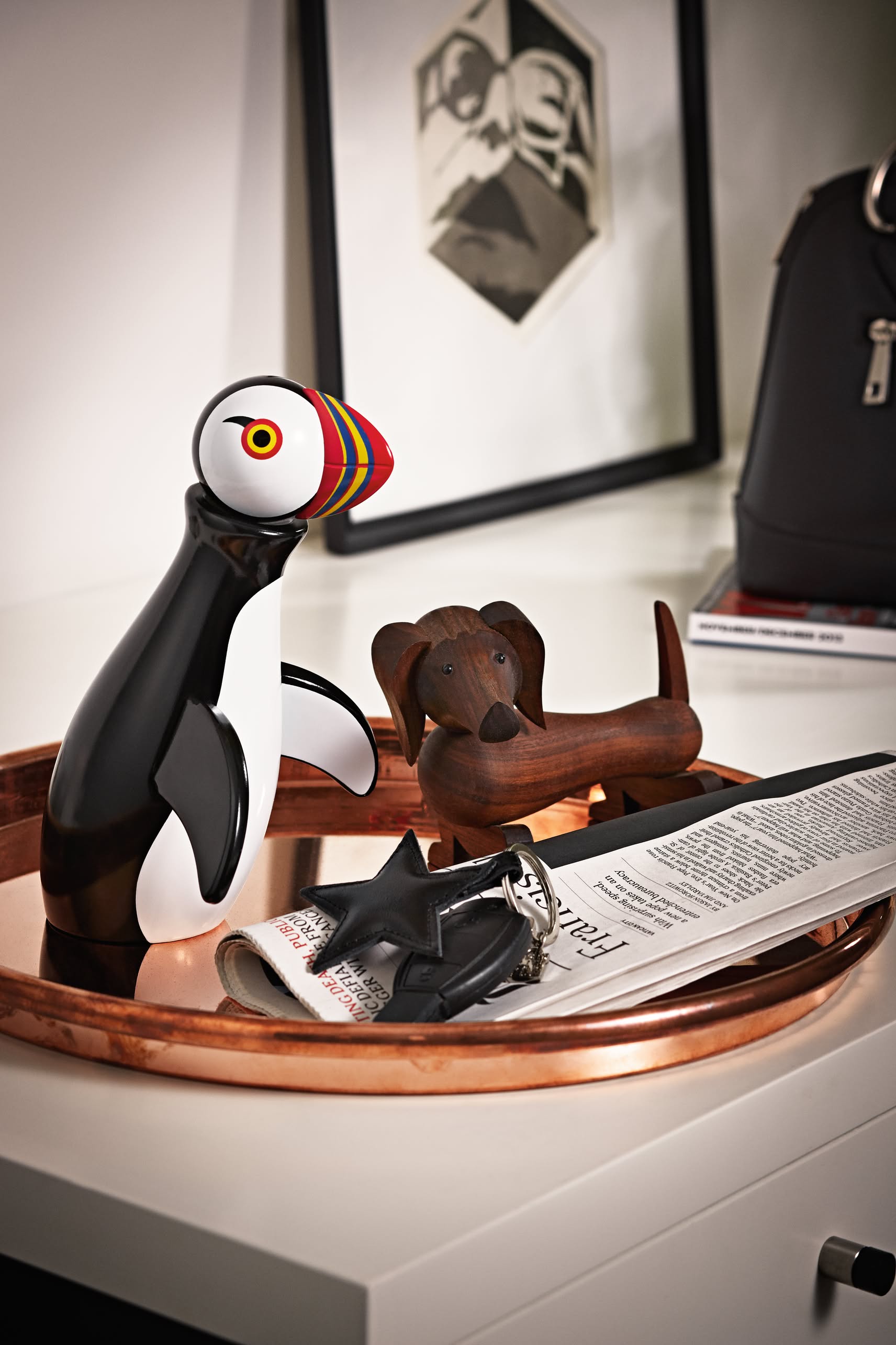 A decorative tray on the table holds a colorful puffin figurine, the Kay Bojesen Dog Small by Kay Bojesen, a folded newspaper, keys, and glasses. In the background are a framed picture and a black bag.