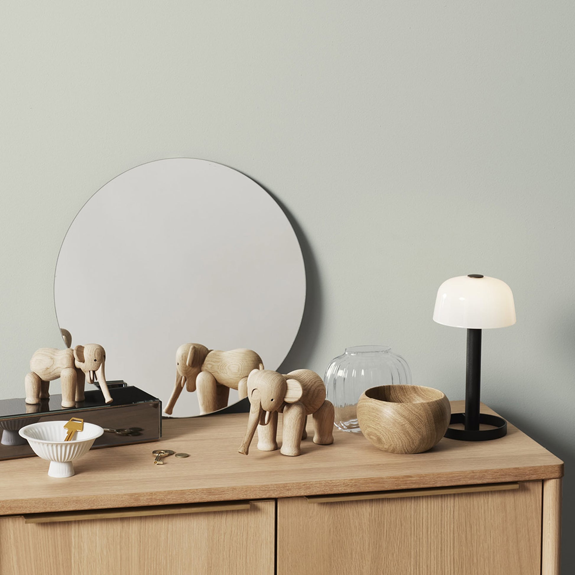 A wooden dresser with a round mirror, two Kay Bojesen Elephant Mini figurines by Kay Bojesen, a glass vase, a wooden bowl, a white table lamp with black base, a white dish with a yellow item, and keys on top.