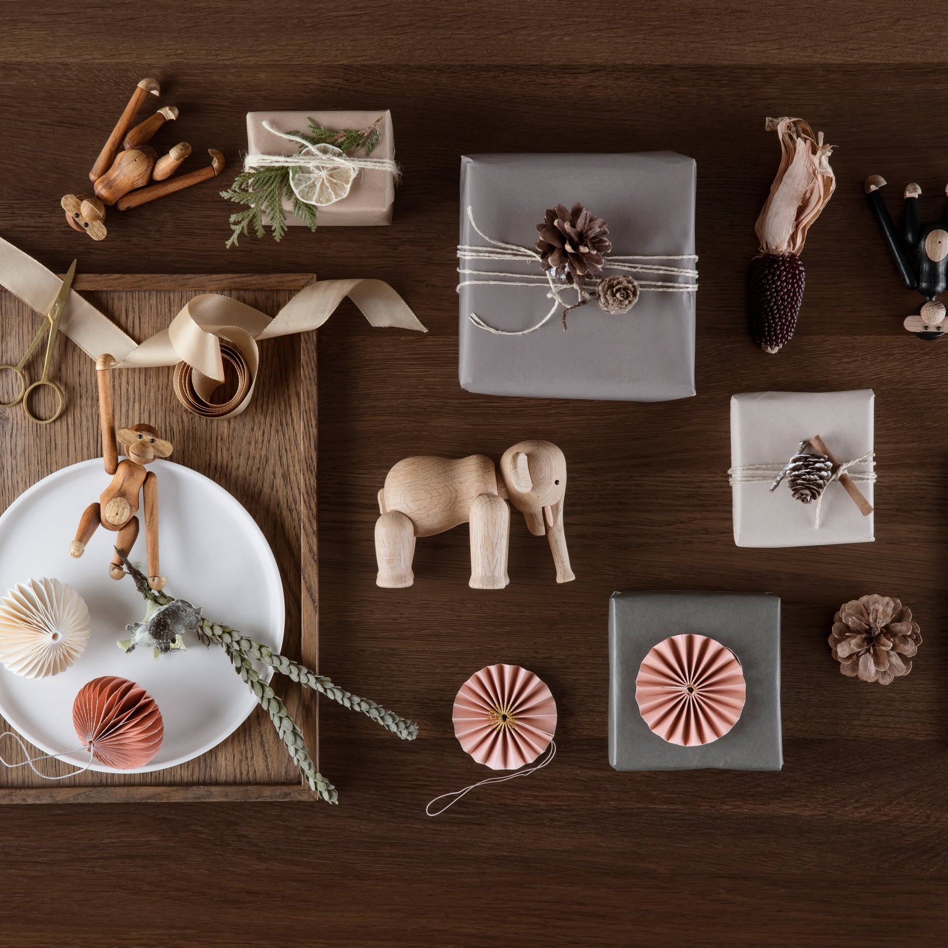 Flat lay of wrapped gifts with natural decorations, wooden toys like the Kay Bojesen Elephant Mini by Kay Bojesen, dried plants, scissors, ribbon, paper ornaments, and pinecones on a wooden table—celebrating Danish design.