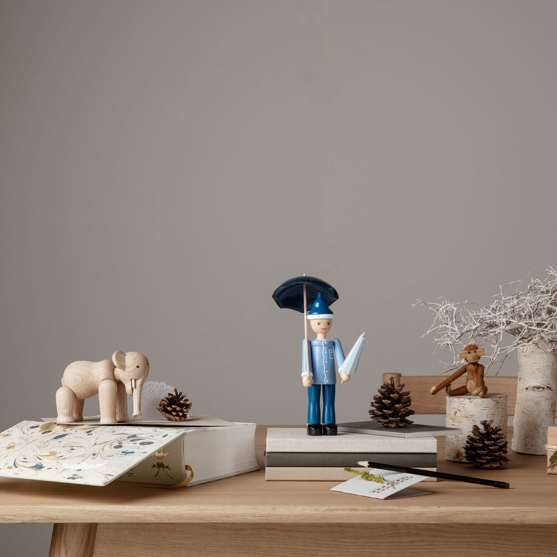 A wooden desk displays stacked books, a Kay Bojesen Elephant Mini by Kay Bojesen exemplifying Danish design, a toy soldier with an umbrella, pinecones, a small wooden monkey, birch decorations, and a dried white branch against a gray wall.