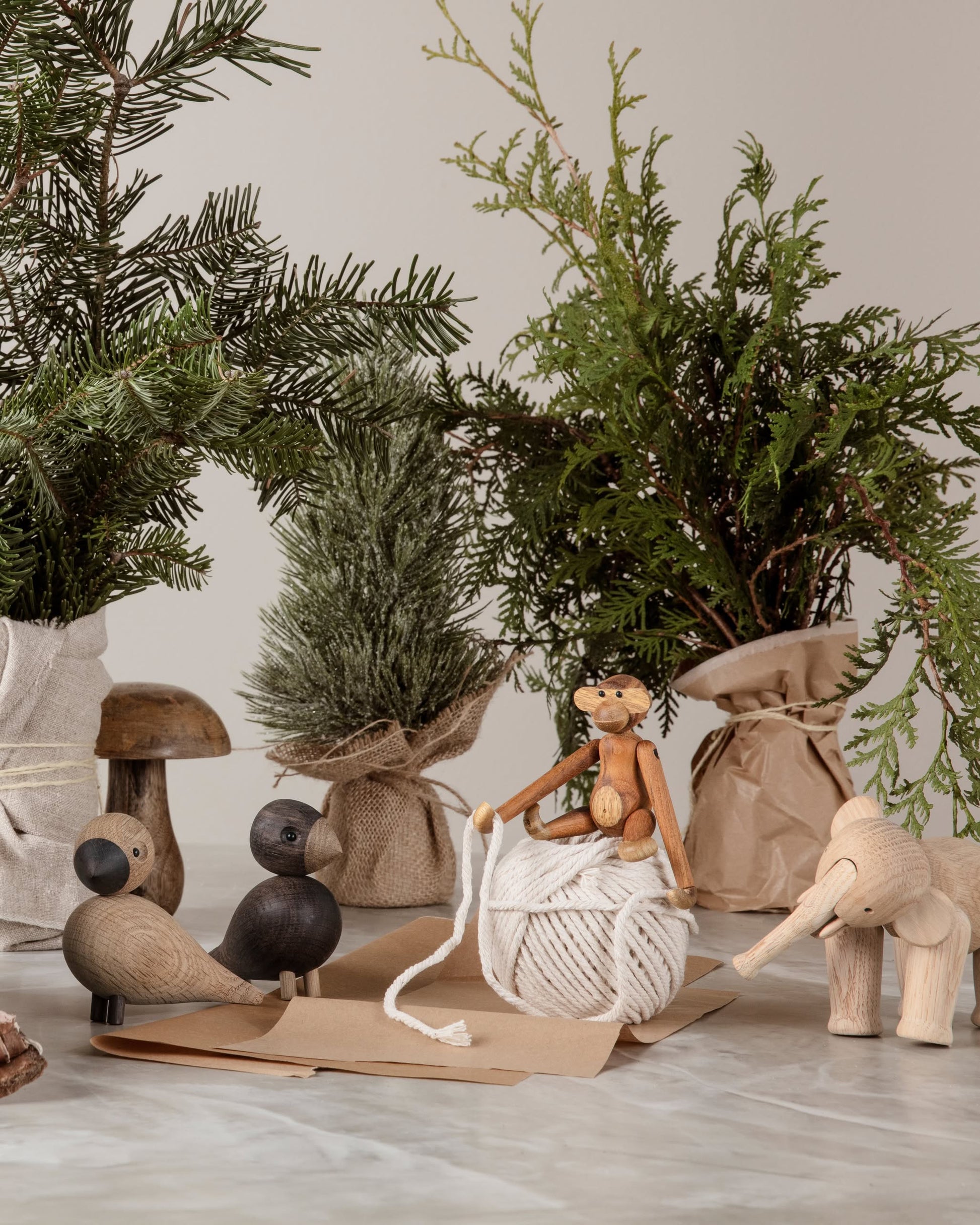 A festive, rustic scene features small potted evergreens, wooden animal figurines, brown paper envelopes, white string, and the Kay Bojesen Monkey Mini by Kay Bojesen perched on the string for a cozy touch.
