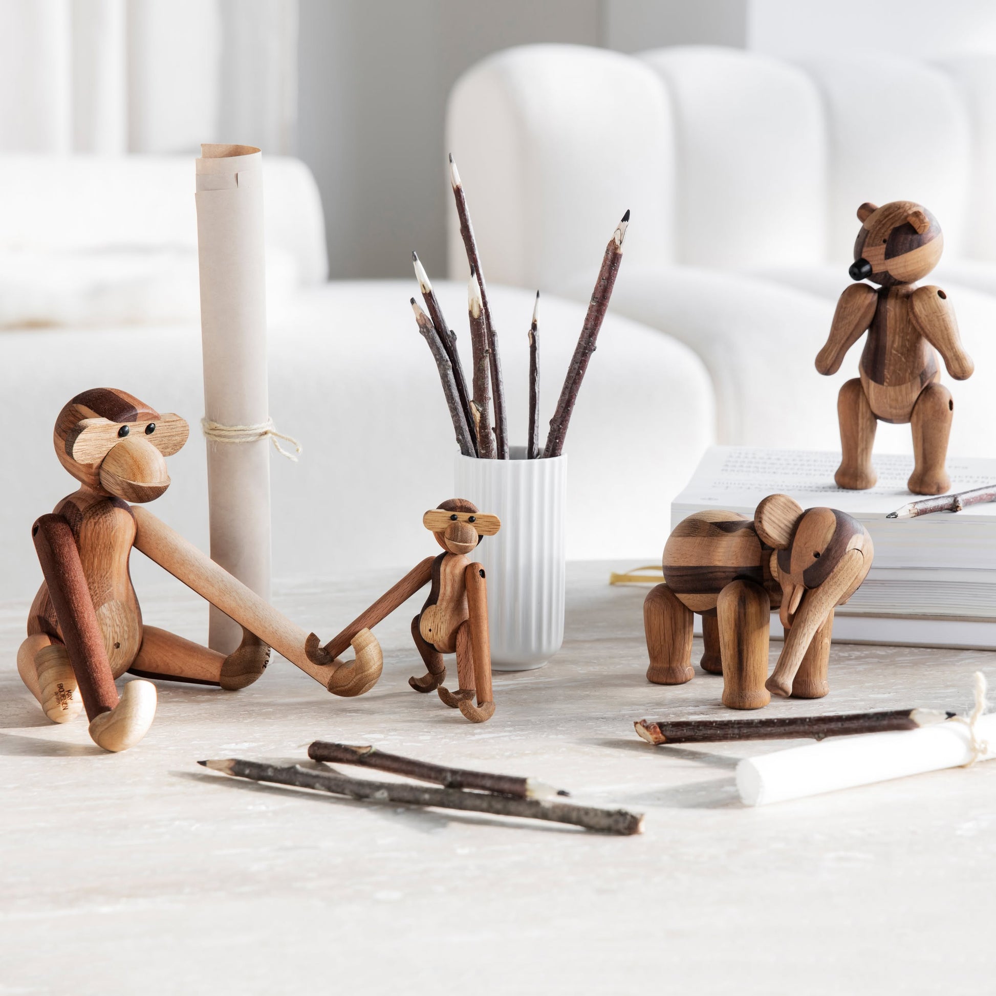 The Kay Bojesen Monkey Mini by Kay Bojesen is displayed on a light table with twig pencils, paper scrolls, and a white pencil holder. A white sofa and books in the background create a charming collectible keepsake setting.