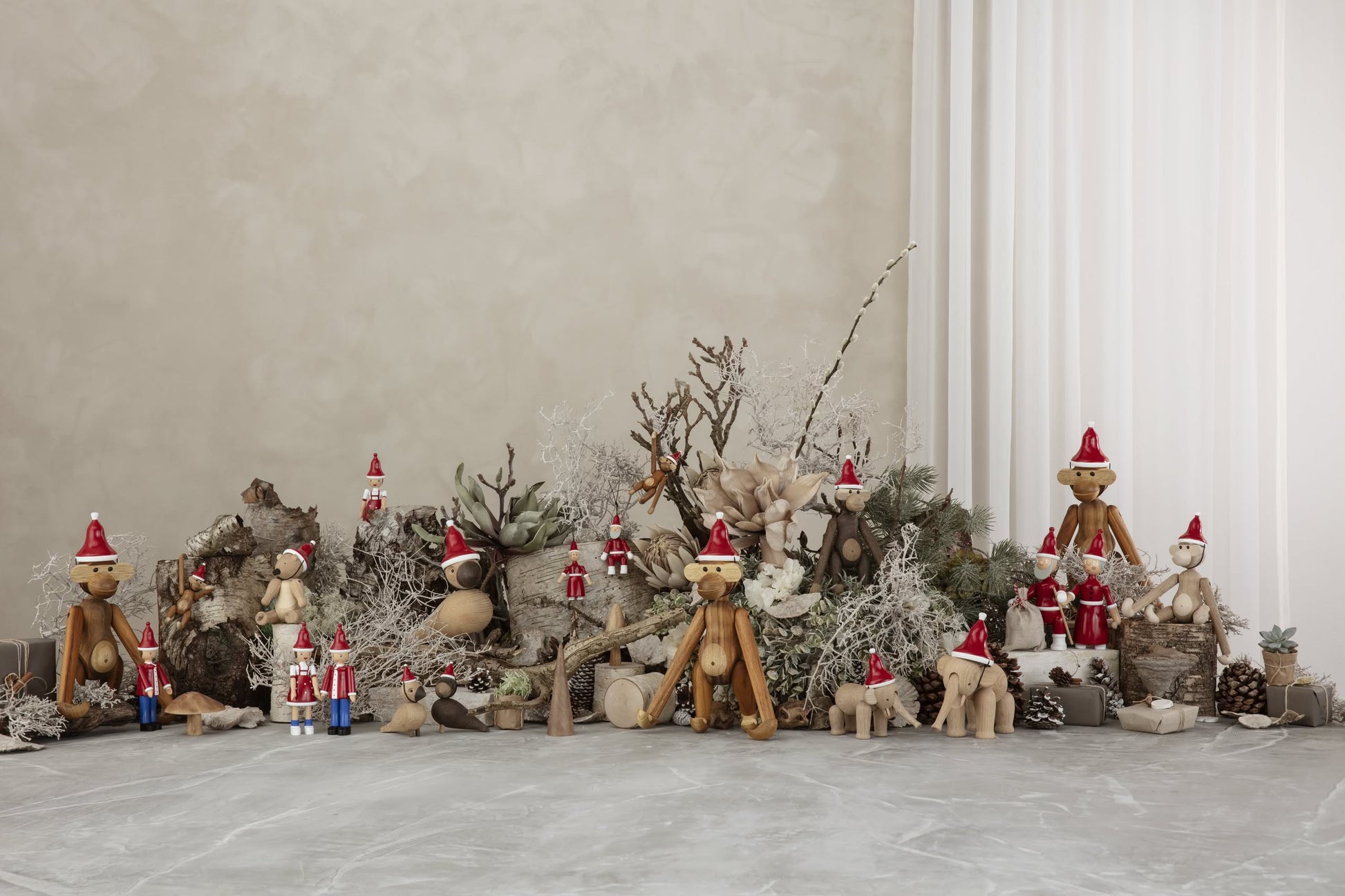 A festive winter scene with collectible wooden animals, featuring the Kay Bojesen Monkey Mini by Kay Bojesen, plus tiny Santa figures arranged among branches, pinecones, and frosted accents against a neutral wall and sheer curtains.