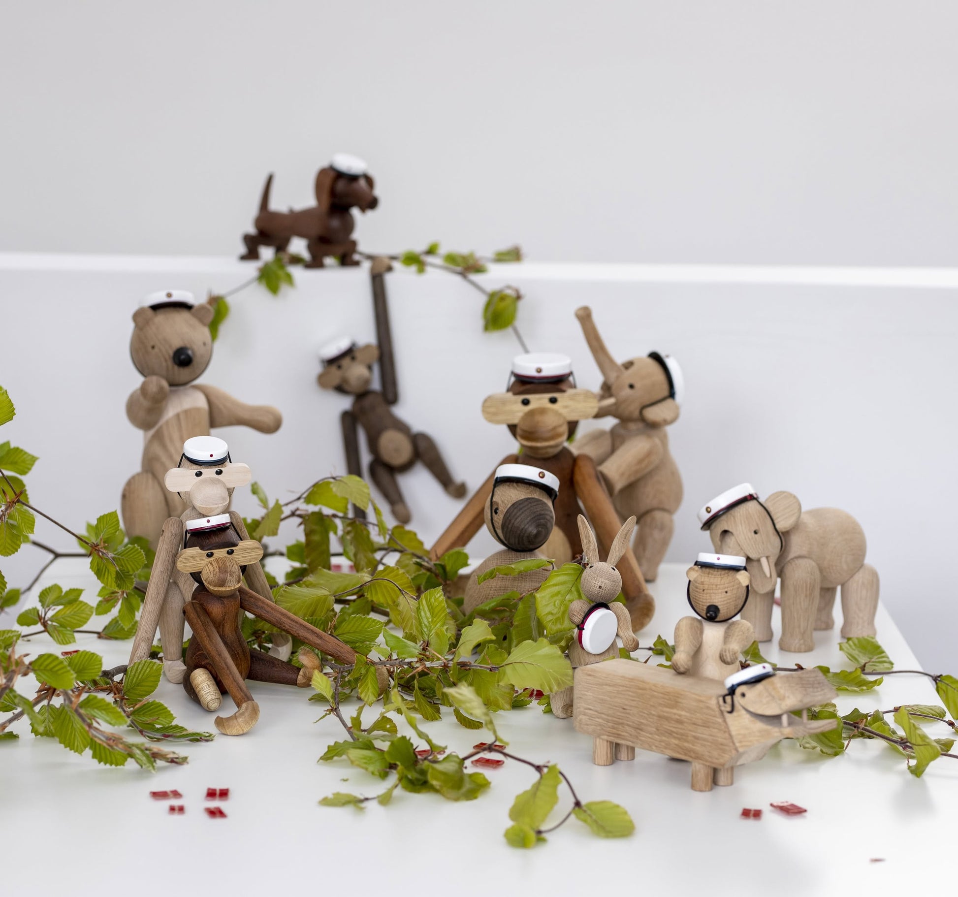 A set of wooden animal figurines, featuring the Kay Bojesen Monkey Mini by Kay Bojesen, each with a tiny white graduation cap, is displayed on a white surface with green branches—ideal as a collectible keepsake or mini display piece.