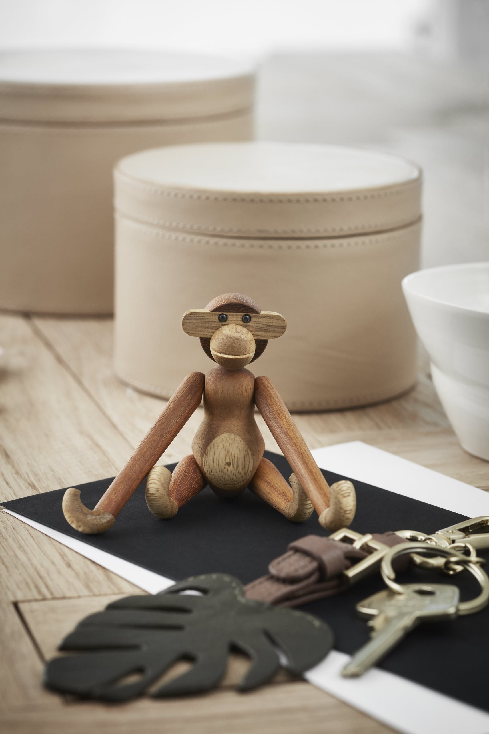 The Kay Bojesen Monkey Mini by Kay Bojesen rests on a black envelope atop a wooden surface, surrounded by beige storage boxes, a white bowl, keys on a keychain, and a black leaf-shaped tag.