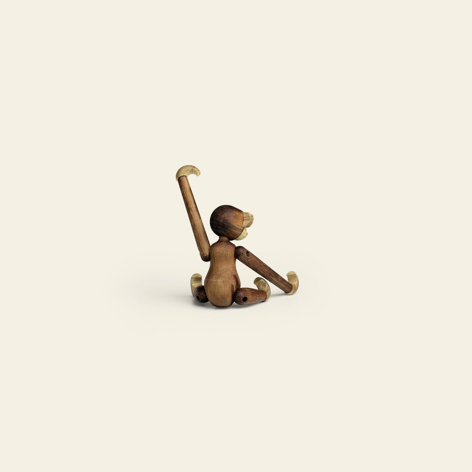The Kay Bojesen Monkey Mini by Kay Bojesen sits on a light, simple background with one arm raised and the other touching the ground, viewed from behind. This charming keepsake stands out for its minimalist design.