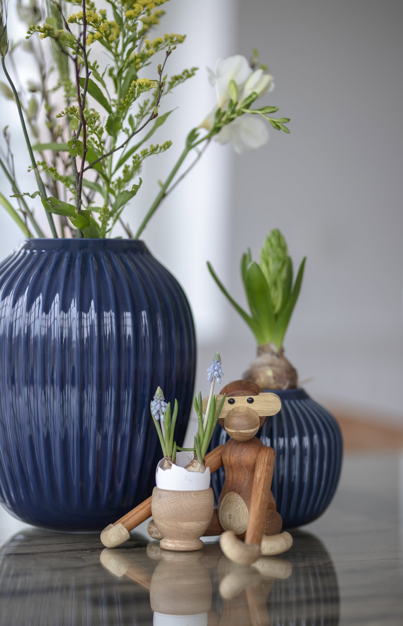 The Kay Bojesen Monkey Mini by Kay Bojesen sits beside two blue vases with flowers and a wooden planter of tiny purple blooms on a reflective surface, its charm enhanced by the softly blurred background.