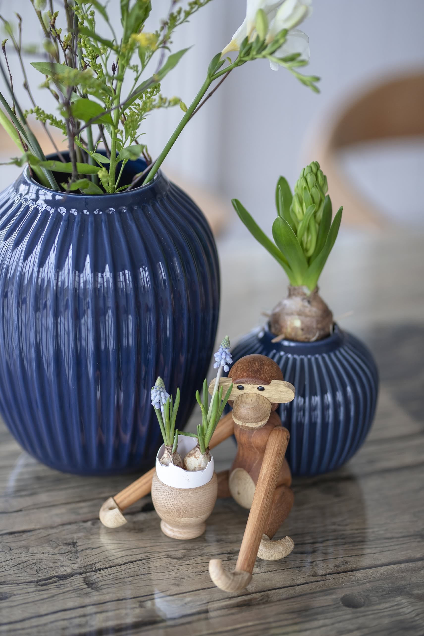A Kay Bojesen Monkey Mini by Kay Bojesen sits beside two blue vases with green plants and flowers on a wooden table, holding a white pot of small blue flowers as a charming collectible keepsake.