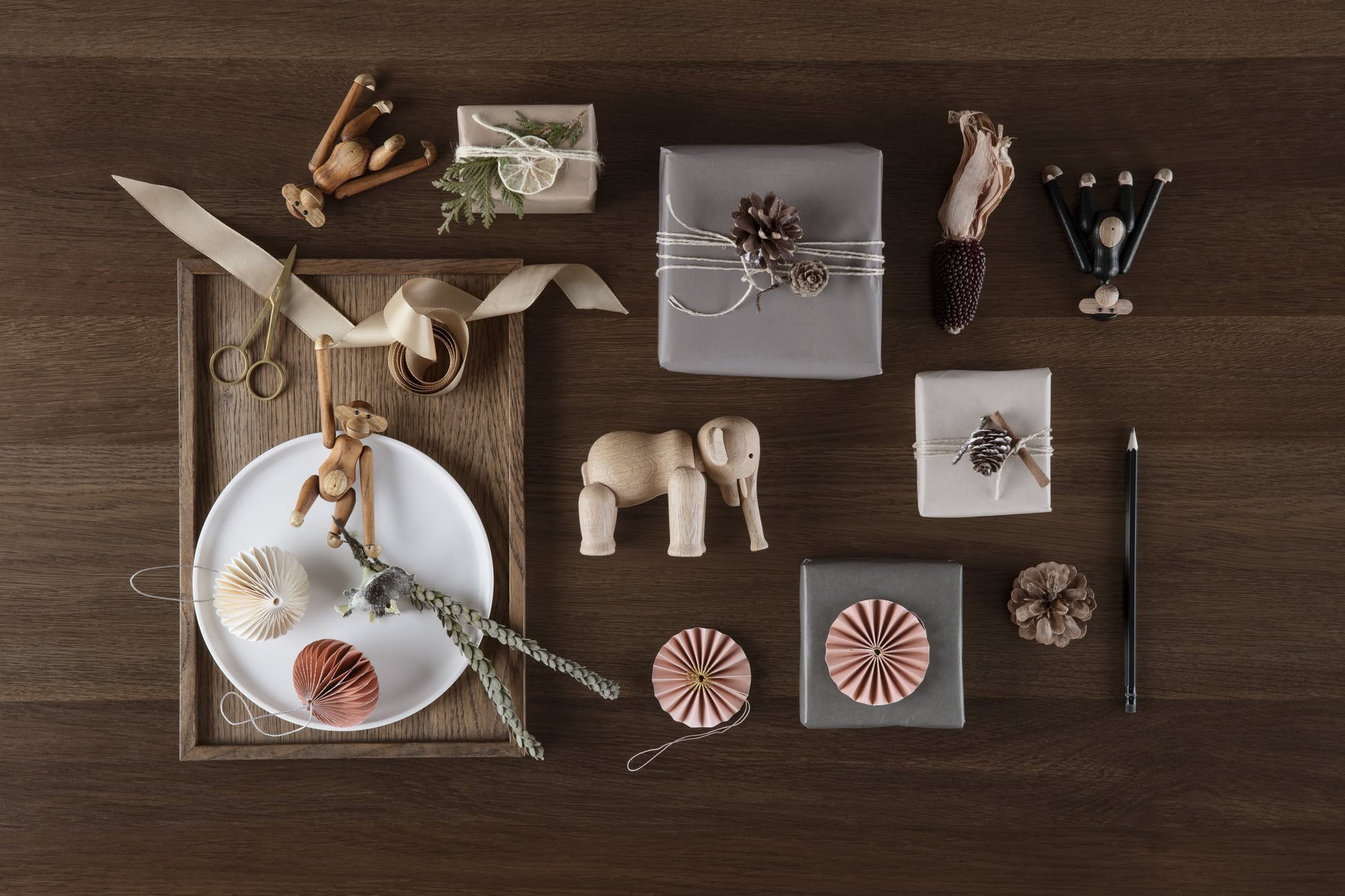 Gift boxes wrapped in neutral paper with natural accents are arranged on a dark wooden surface, accompanied by the Kay Bojesen Monkey Mini from Kay Bojesen, as well as a plate, ribbon, pencil, and small ornaments.