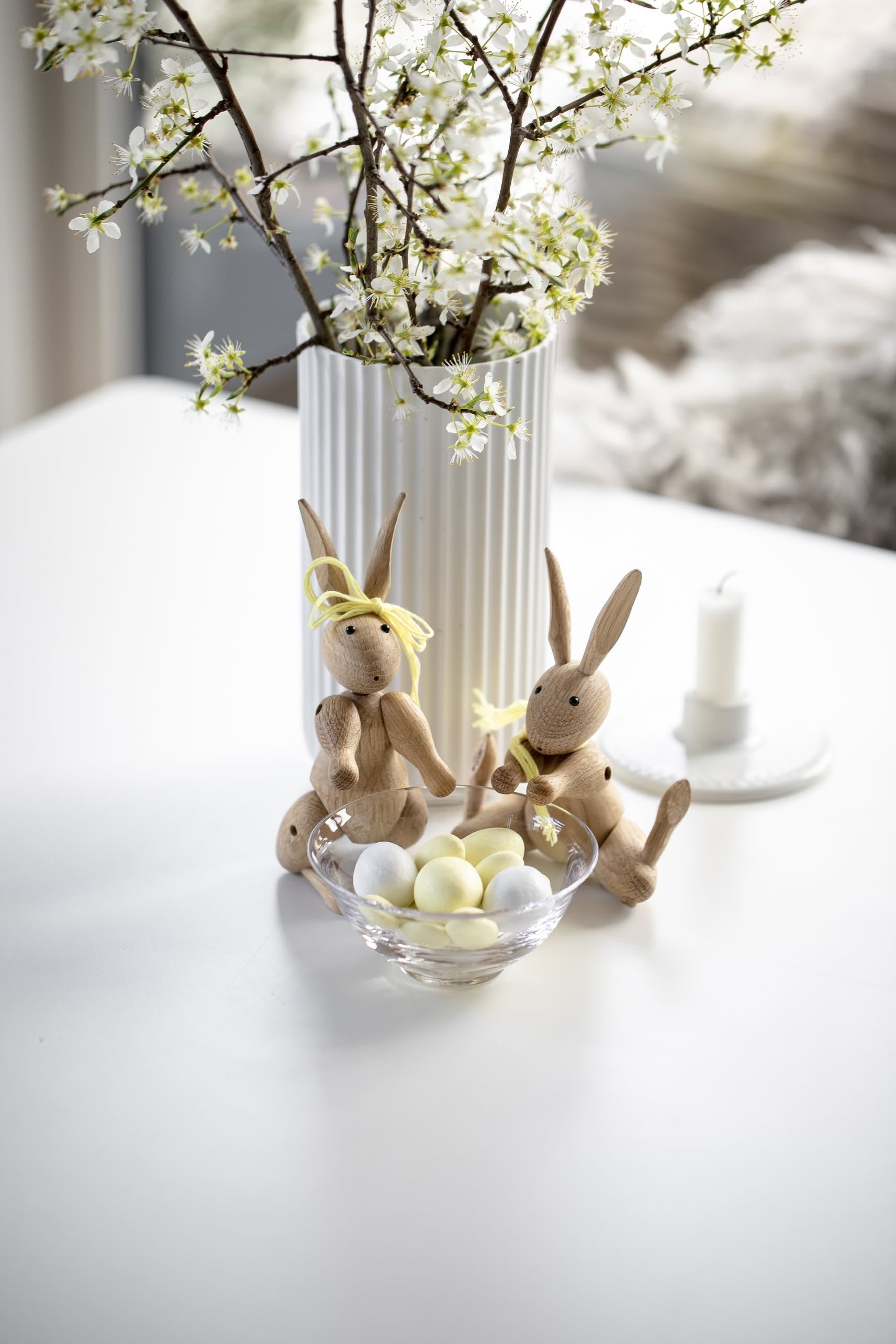 Two Kay Bojesen Rabbit Mini wooden toys by Kay Bojesen with yellow ribbons sit beside a glass bowl of pastel eggs on a white table, with a white vase of blossoming branches and a white candle in the background.