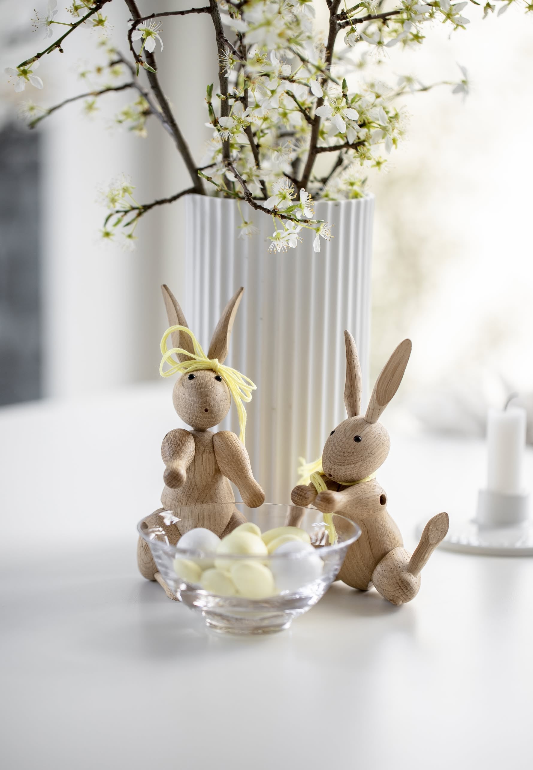 Two whimsical Kay Bojesen Rabbit Mini figurines by Kay Bojesen sit beside a glass bowl of pastel eggs on a white table, framed by blossoming branches. One wears a yellow ribbon; both hold small flowers, echoing Danish craftsmanship.