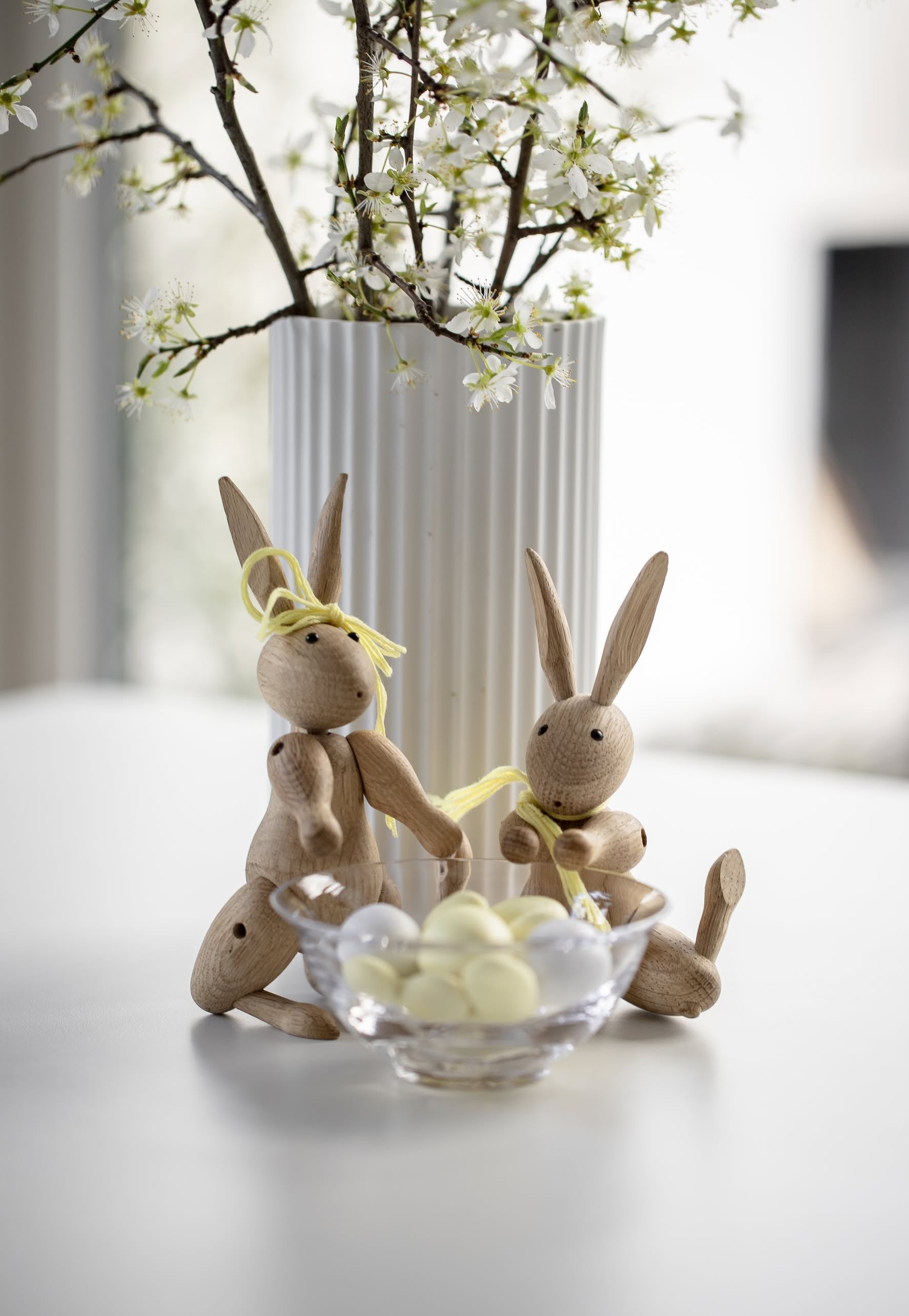 Two Kay Bojesen Rabbit Mini wooden toys with yellow ribbons from Kay Bojesen sit beside a glass bowl of white candies and a white vase of blooming branches on a white table.