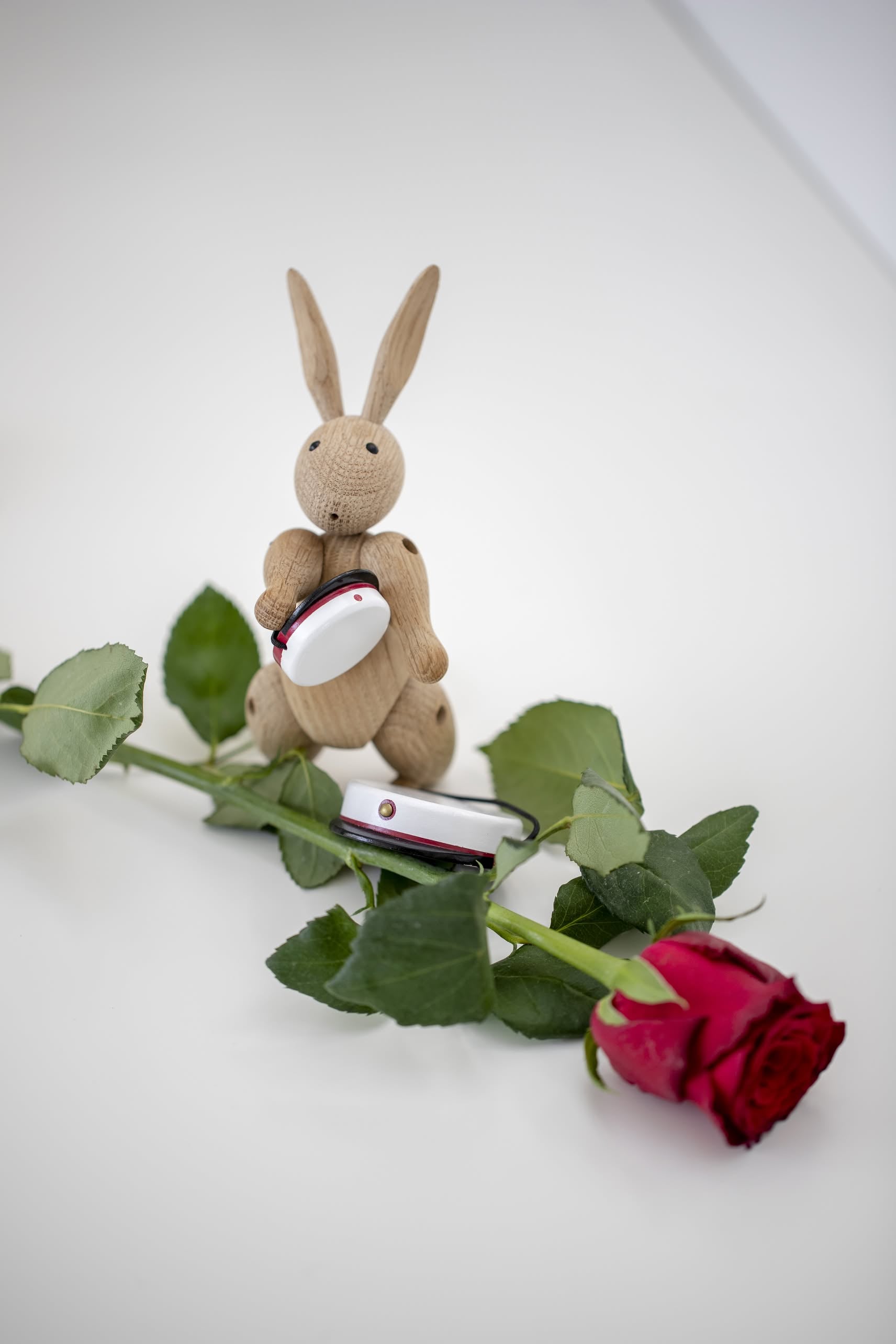 The Kay Bojesen Rabbit Mini by Kay Bojesen, a delightful piece of Danish design, stands beside a small white suitcase, a red rose, and another petite suitcase on a white surface.