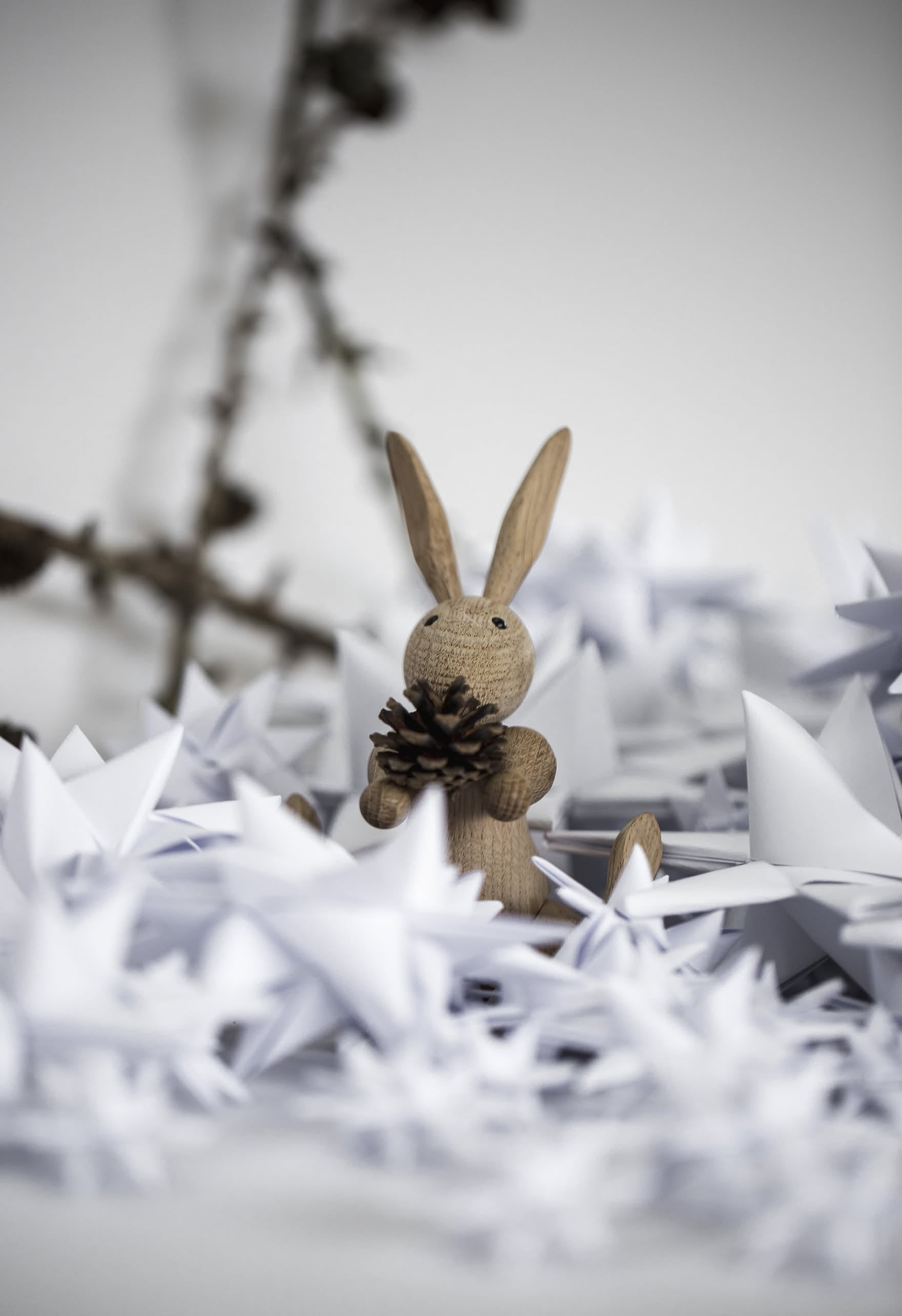 The Kay Bojesen Rabbit Mini by Kay Bojesen, inspired by classic Danish wooden toys, holds a pine cone among white paper stars, with a blurred branch in the background.