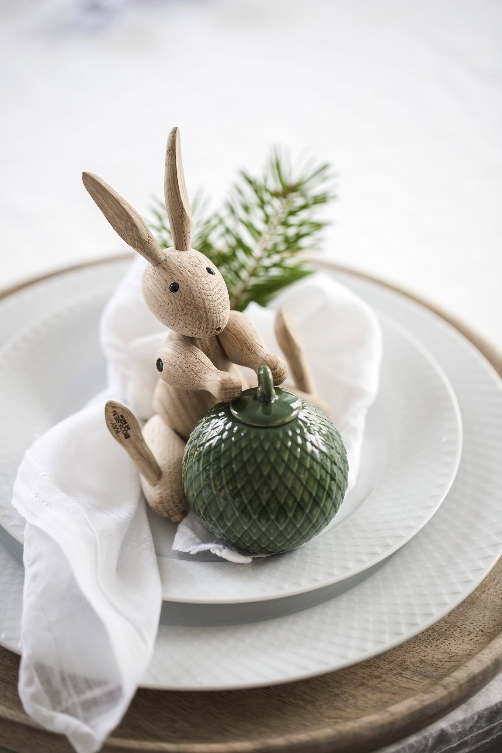The Kay Bojesen Rabbit Mini by Kay Bojesen holds a textured green jar atop stacked white plates with a napkin and pine branch, adding a touch of Danish craftsmanship to your table decor.