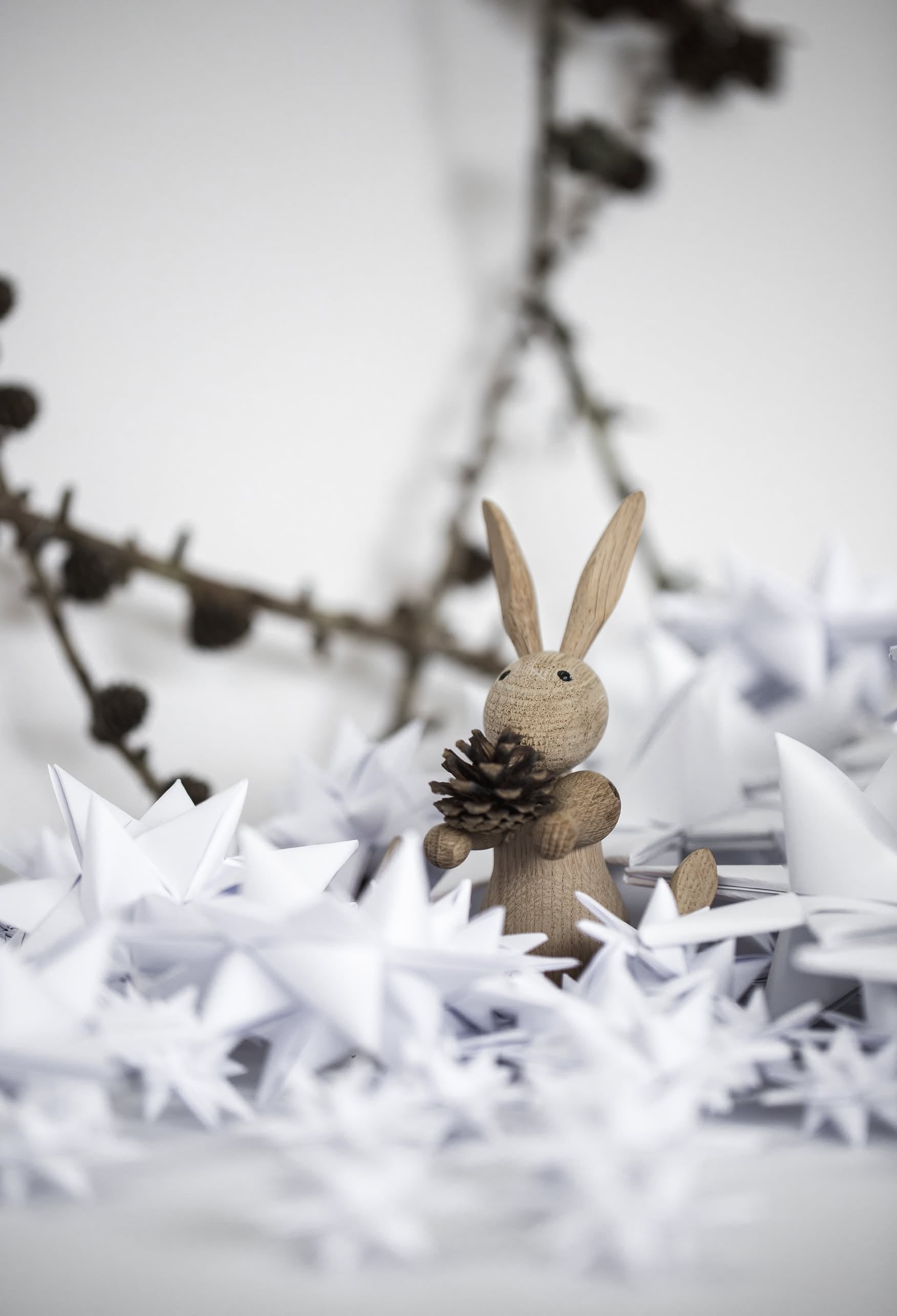 The Kay Bojesen Rabbit Mini by Kay Bojesen holds a pine cone among white origami stars, celebrating Danish craftsmanship, with a blurred branch in the background.