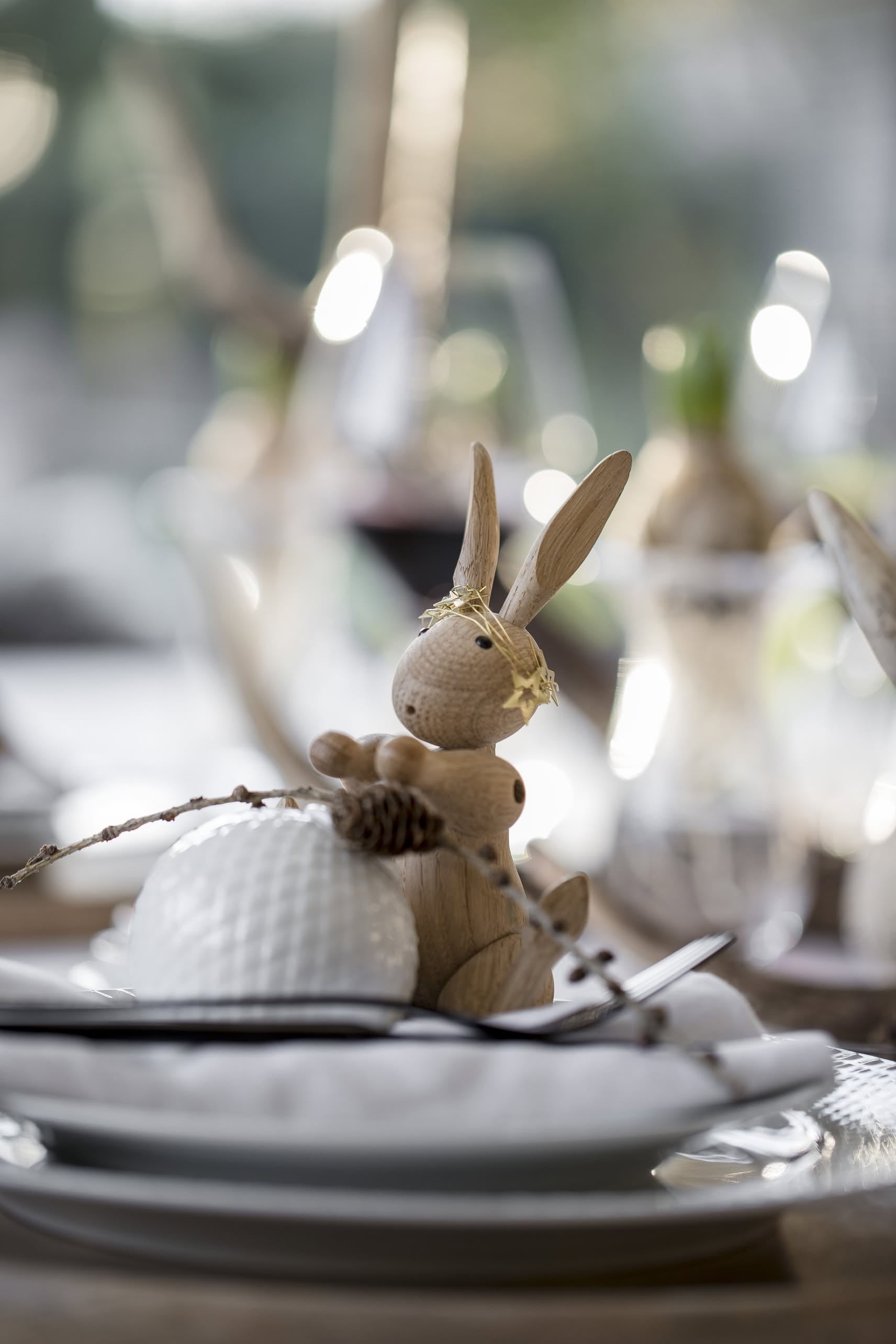 The Kay Bojesen Rabbit Mini by Kay Bojesen, topped with a gold crown, sits on a white plate amid Danish craftsmanship, cutlery, and a pinecone, with blurred glassware and greenery in the background.