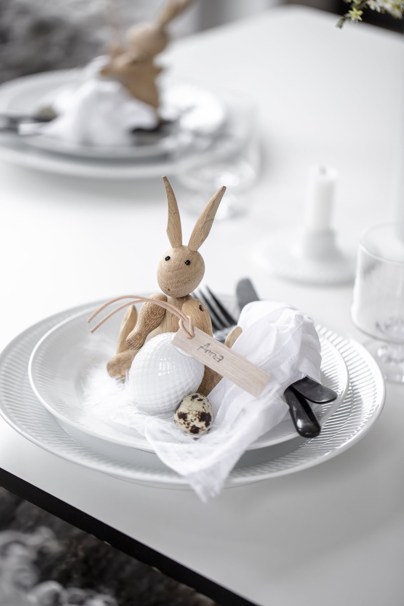 A white plate is arranged with a Kay Bojesen Rabbit Mini by Kay Bojesen, a textured white egg, a small spotted egg, cutlery wrapped in a napkin, and a name tag for an elegant Easter table setting that highlights Danish craftsmanship.