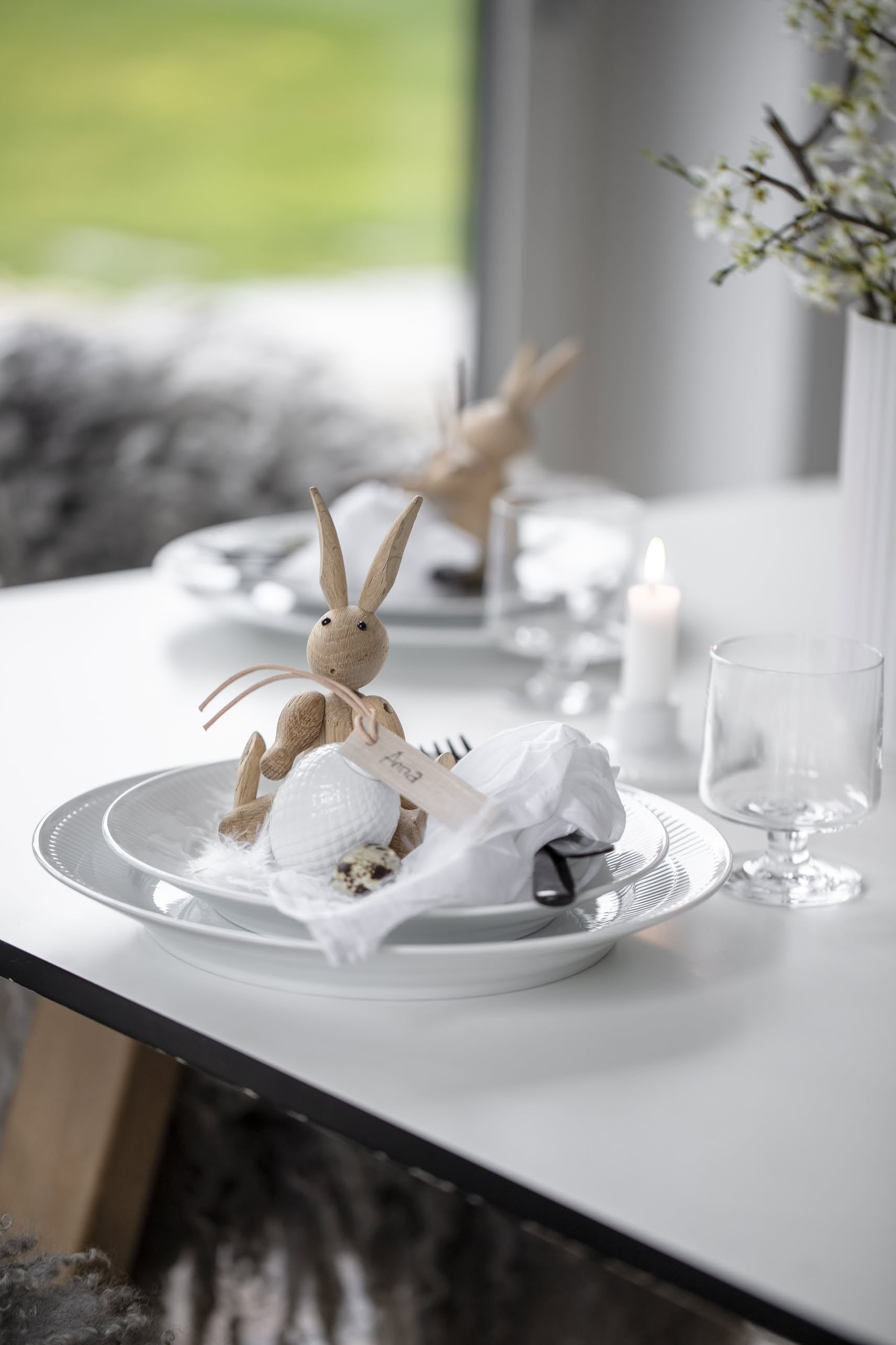 A white table is set with a white plate, glass, napkin, and the Kay Bojesen Rabbit Mini by Kay Bojesen. A lit candle and a vase of white flowers highlight Danish craftsmanship, with another similar place setting in the background.