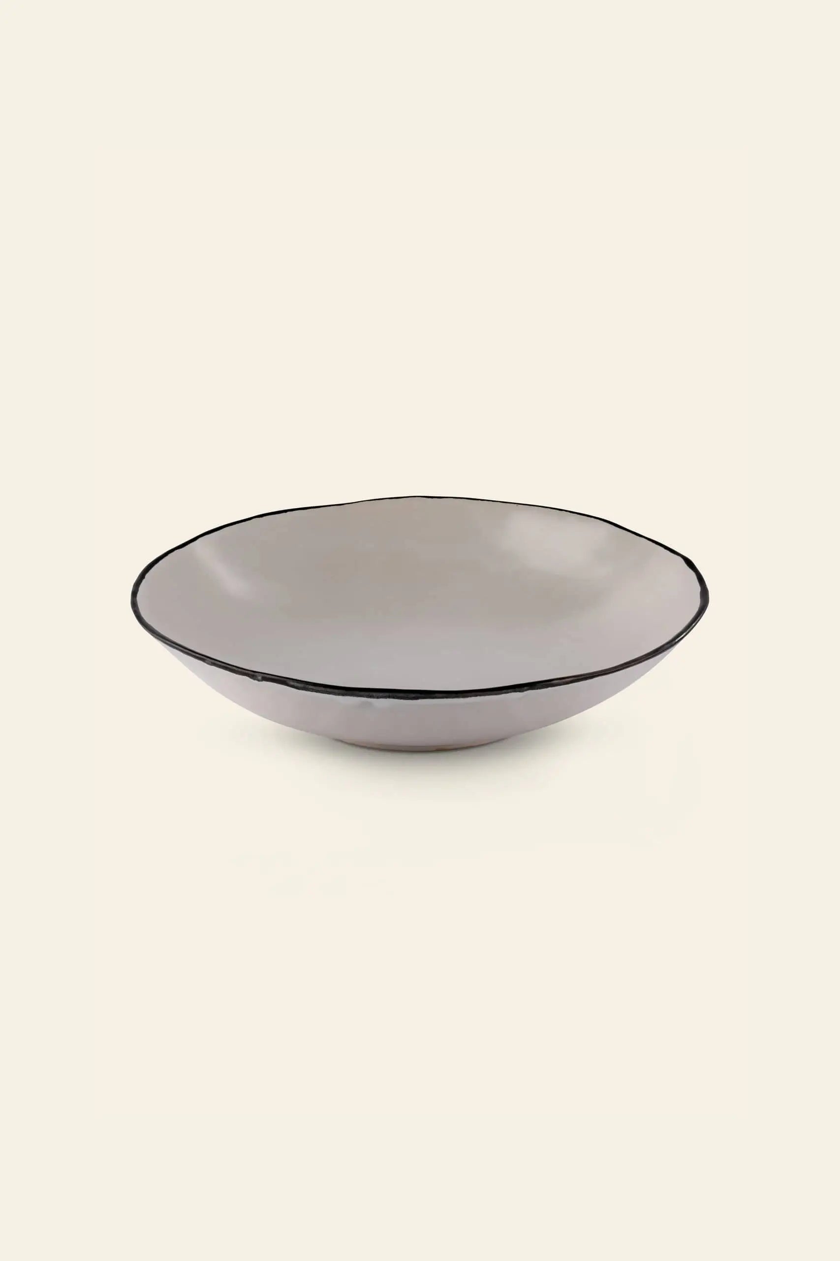 The Kra Sanctuary Andaman Free-Form Pasta Bowl is a shallow, round beige ceramic bowl with a slightly irregular rim and dark edge, hand-painted glaze, showcased against a plain light backdrop.