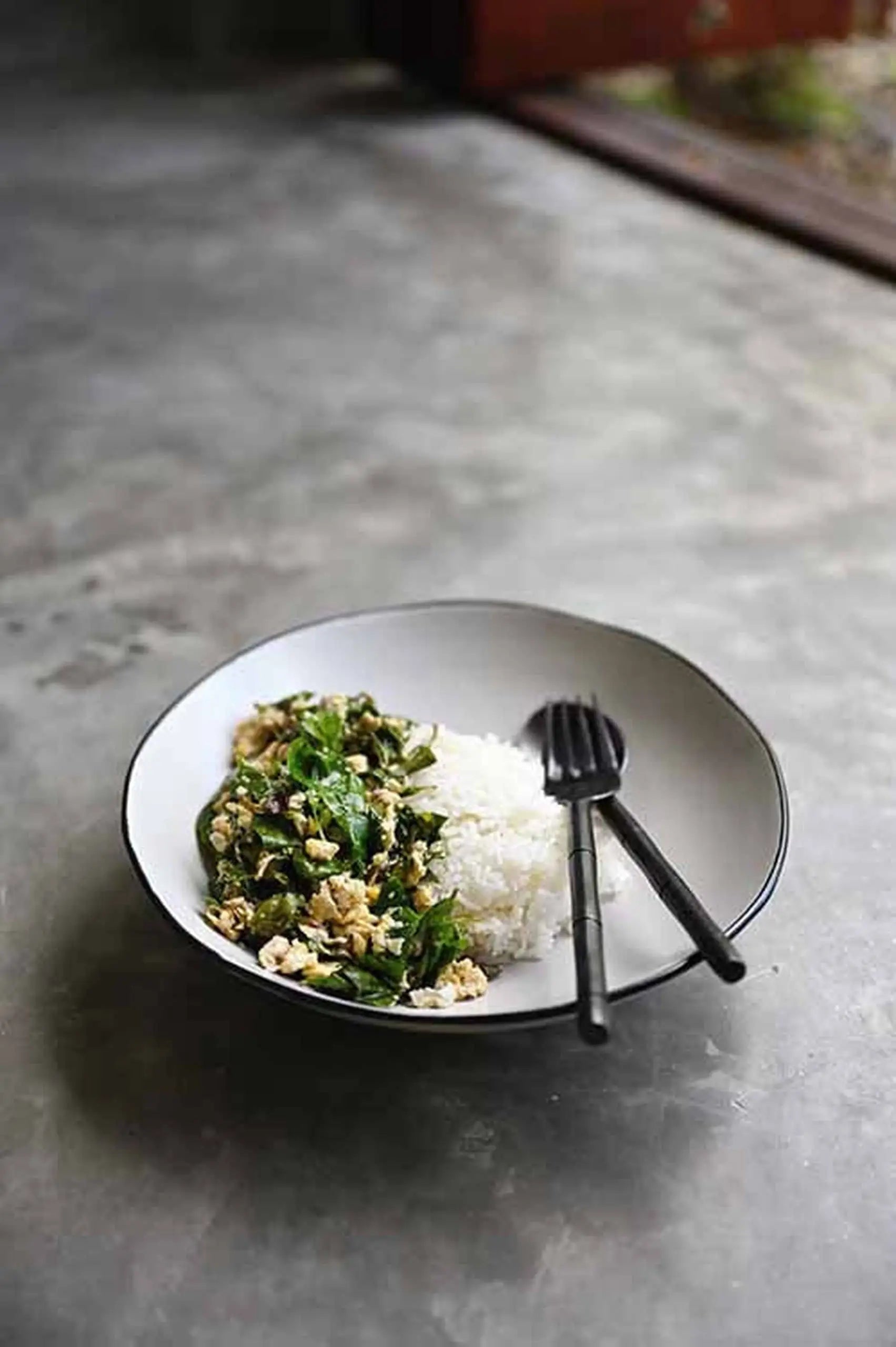 The Kra Sanctuary Andaman Free-Form Pasta Bowl in white by Kra Sanctuary holds steamed rice, stir-fried greens, and tofu on a gray surface. A black fork and spoon complete this elegant island-inspired dining setting.