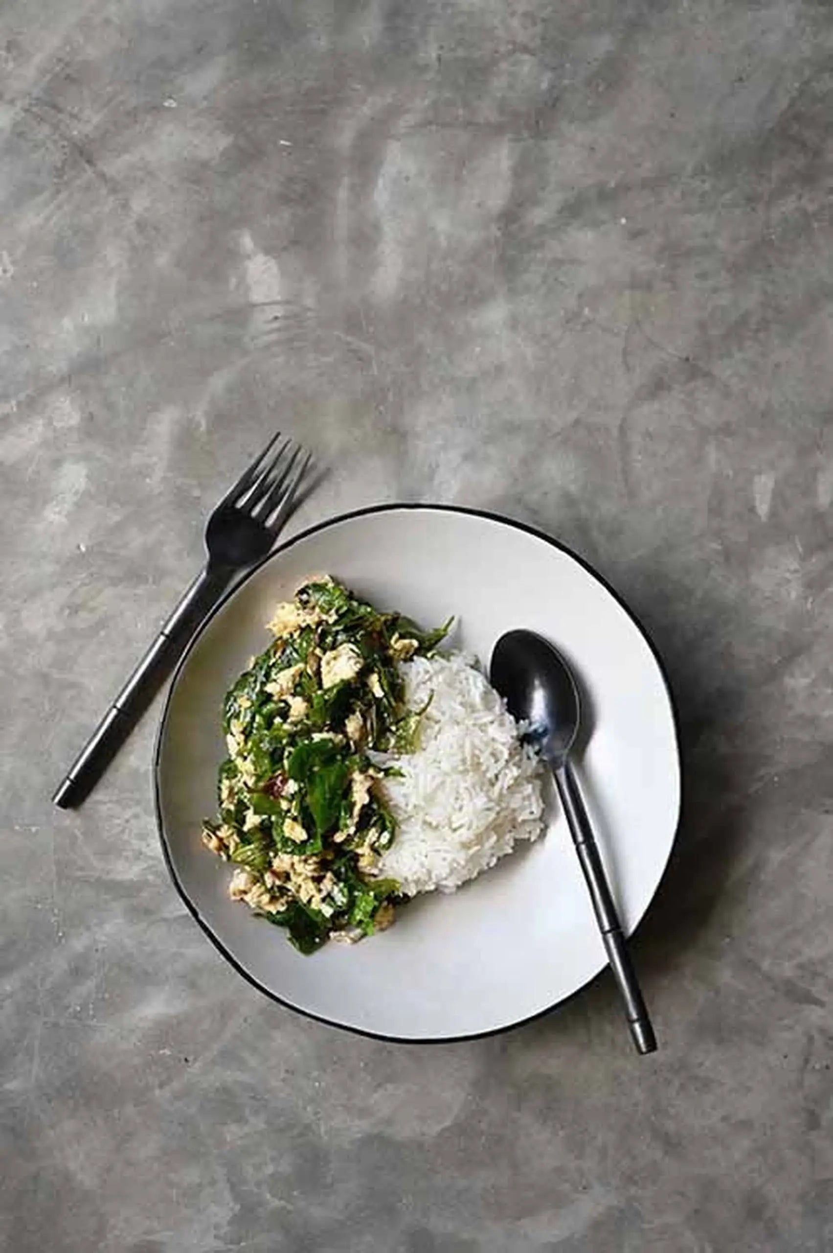 White rice and mixed green vegetable stir-fry are served in the Kra Sanctuary Andaman Free-Form Pasta Bowl by Kra Sanctuary, set on a gray surface with a fork and spoon on either side.