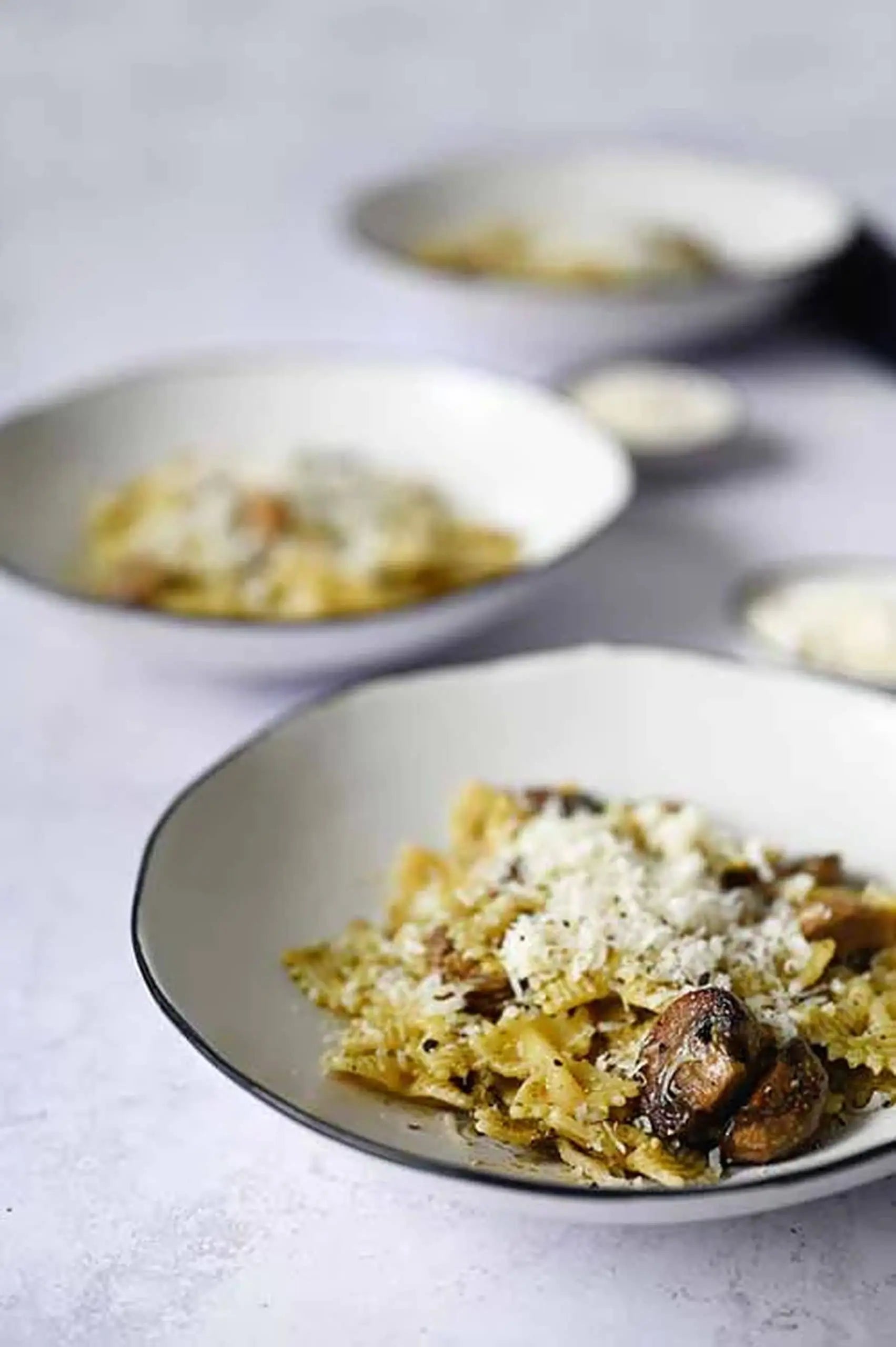 A Kra Sanctuary Andaman Free-Form Pasta Bowl with hand-painted glaze holds farfalle, mushrooms, and cheese on a light surface, creating an island vibe. Two more identical bowls of the same dish are blurred in the background.