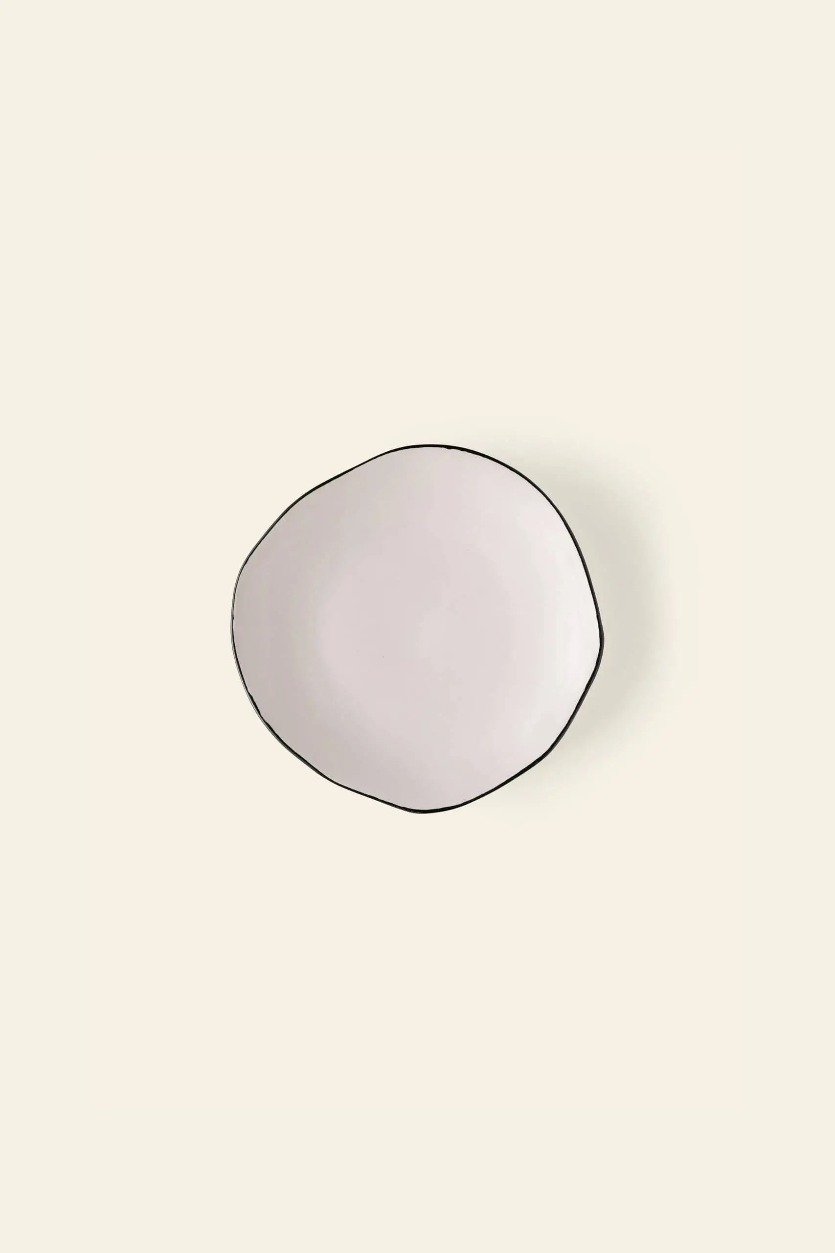 The Kra Sanctuary Andaman Free-Form Side Plate by Kra Sanctuary is a round, off-white ceramic plate with an irregular shape and thin dark rim, reflecting relaxed island elegance against a light beige background.