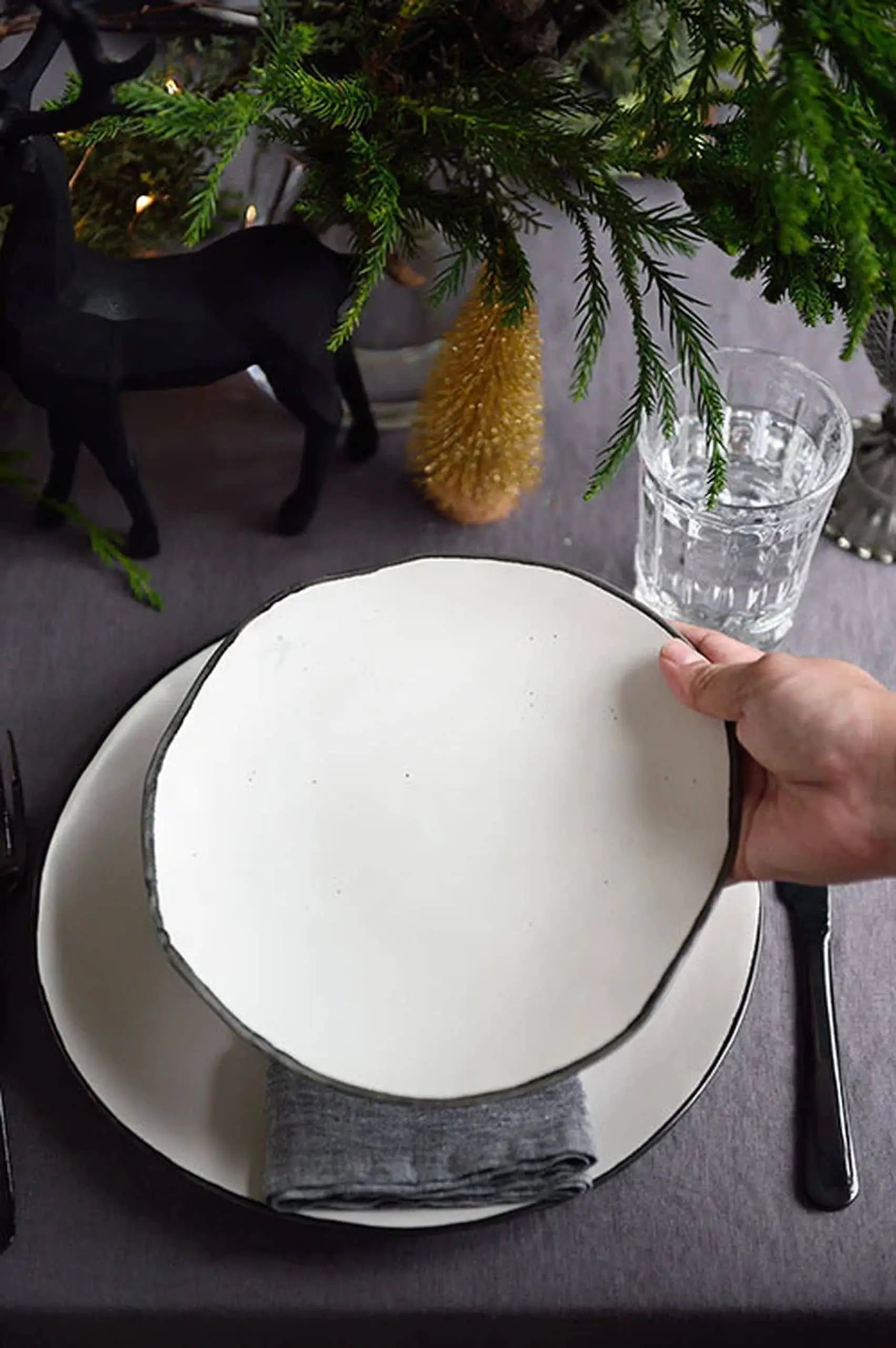 A hand places the Kra Sanctuary Andaman Free-Form Side Plate, a round white ceramic piece with a rustic edge, atop another plate on a gray napkin. The table features greenery, a black reindeer figurine, and a glass of water for an elevated look.
