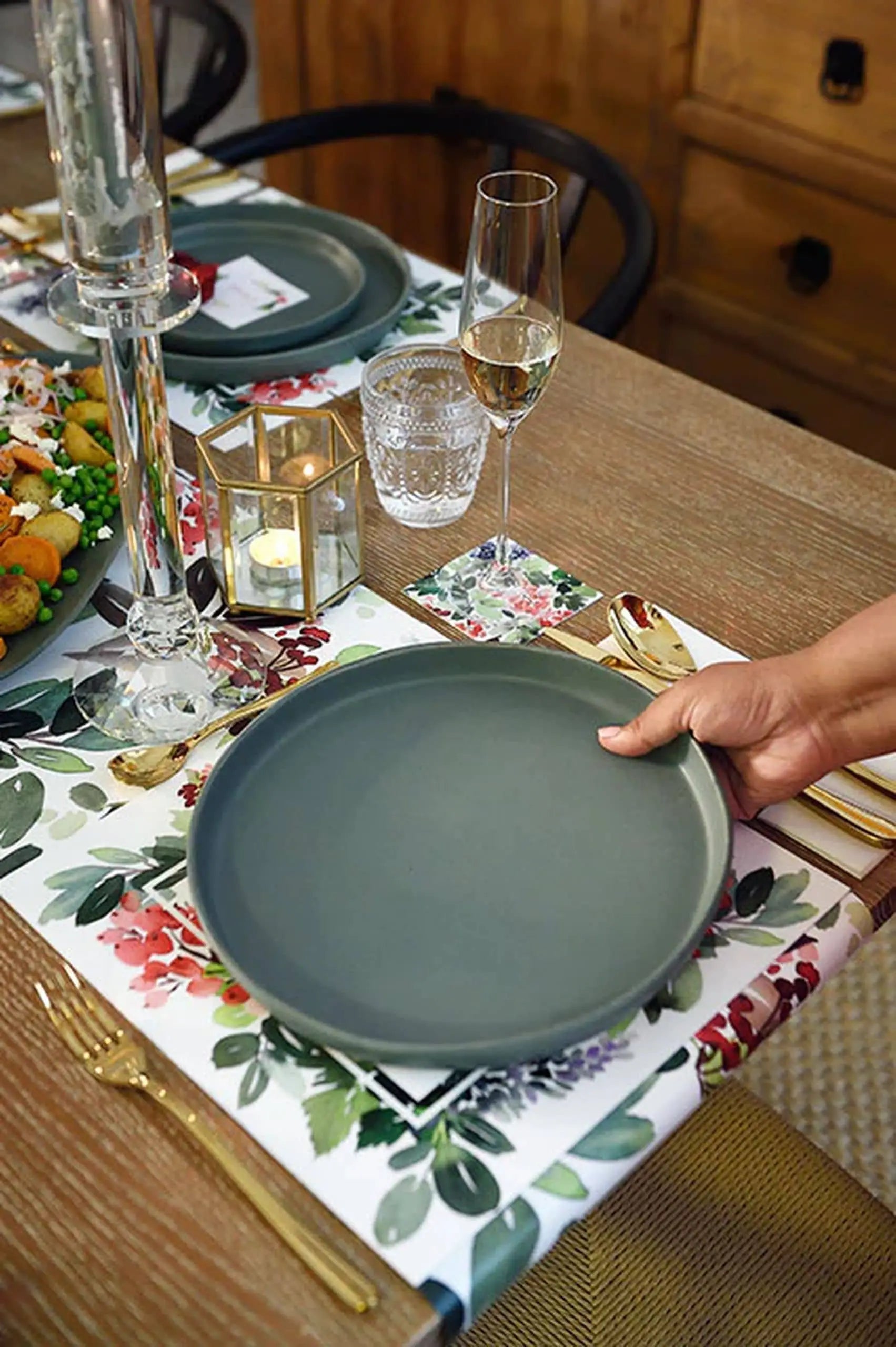 A hand sets a Kra Sanctuary Karst Flat Dinner Plate, in gray, beside gold cutlery, wine and water glasses, and a lit candle on a wooden table, creating rustic charm for a memorable meal.