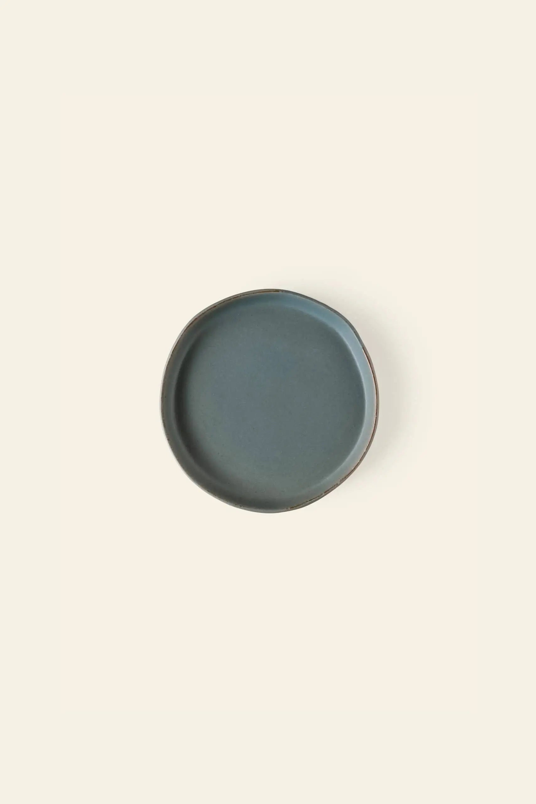 The Kra Sanctuary Karst Flat Side Plate by Kra Sanctuary is a small, round, matte blue ceramic plate with a slightly irregular edge, shown from above on a plain light beige background.