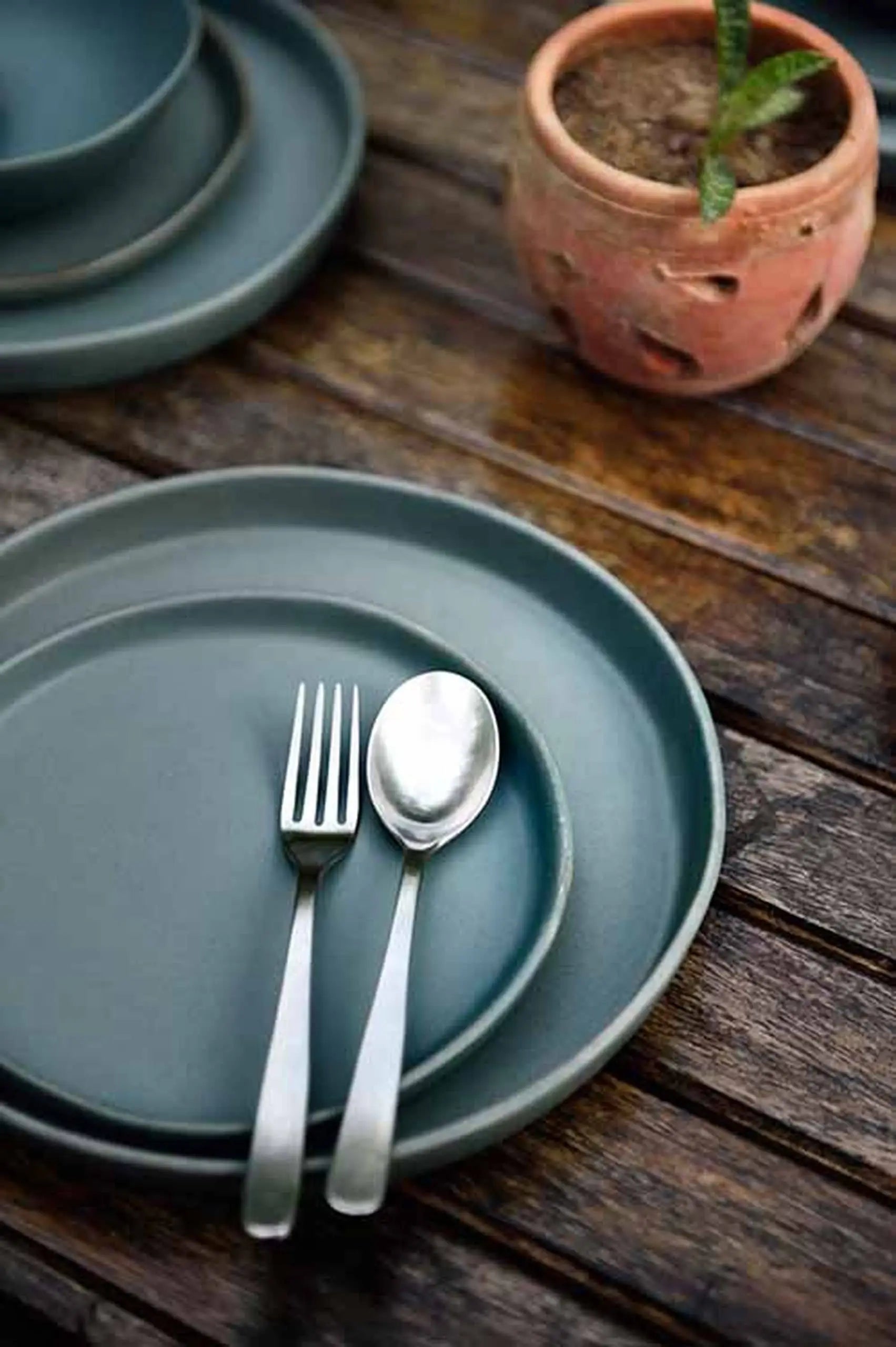The Kra Sanctuary Karst Flat Side Plate is styled with a fork and spoon on a rustic wooden table beside a clay pot with a small plant, adding charm to your dining setting.