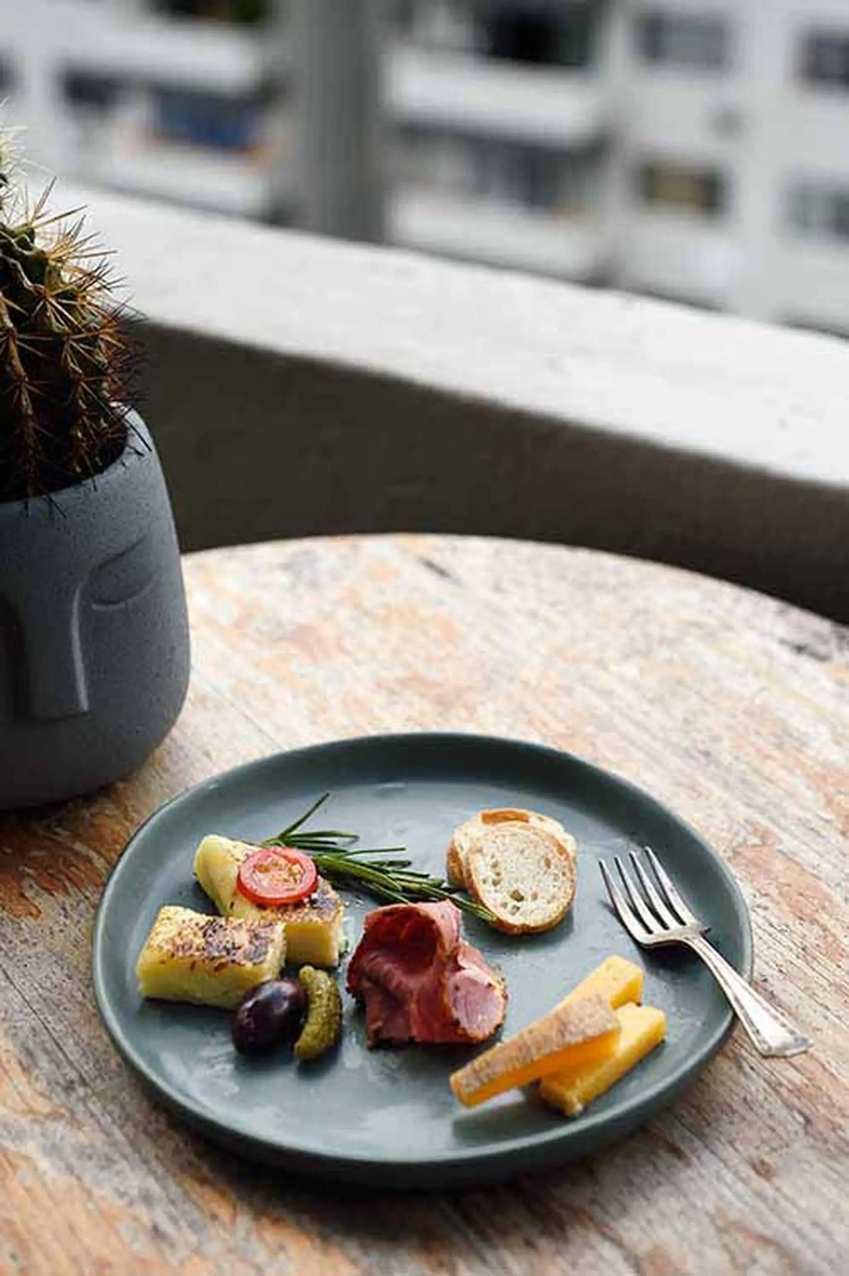 A Kra Sanctuary Karst Flat Side Plate holds an assortment of meat, cheese, olives, and bread on a rustic table beside a potted cactus, adding charm to the scene with a blurred cityscape in the background.