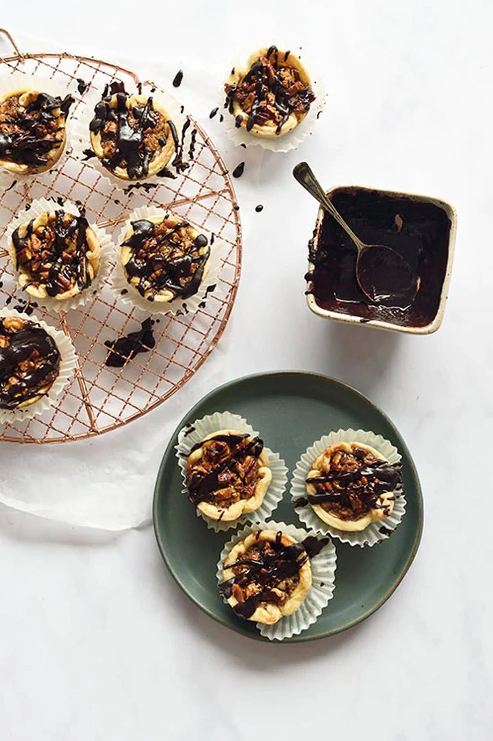Mini tarts with nut and chocolate topping are arranged on a wire rack and a Kra Sanctuary Karst Flat Side Plate, adding rustic charm to the scene, with a small bowl of chocolate sauce and spoon beside them on a white surface.