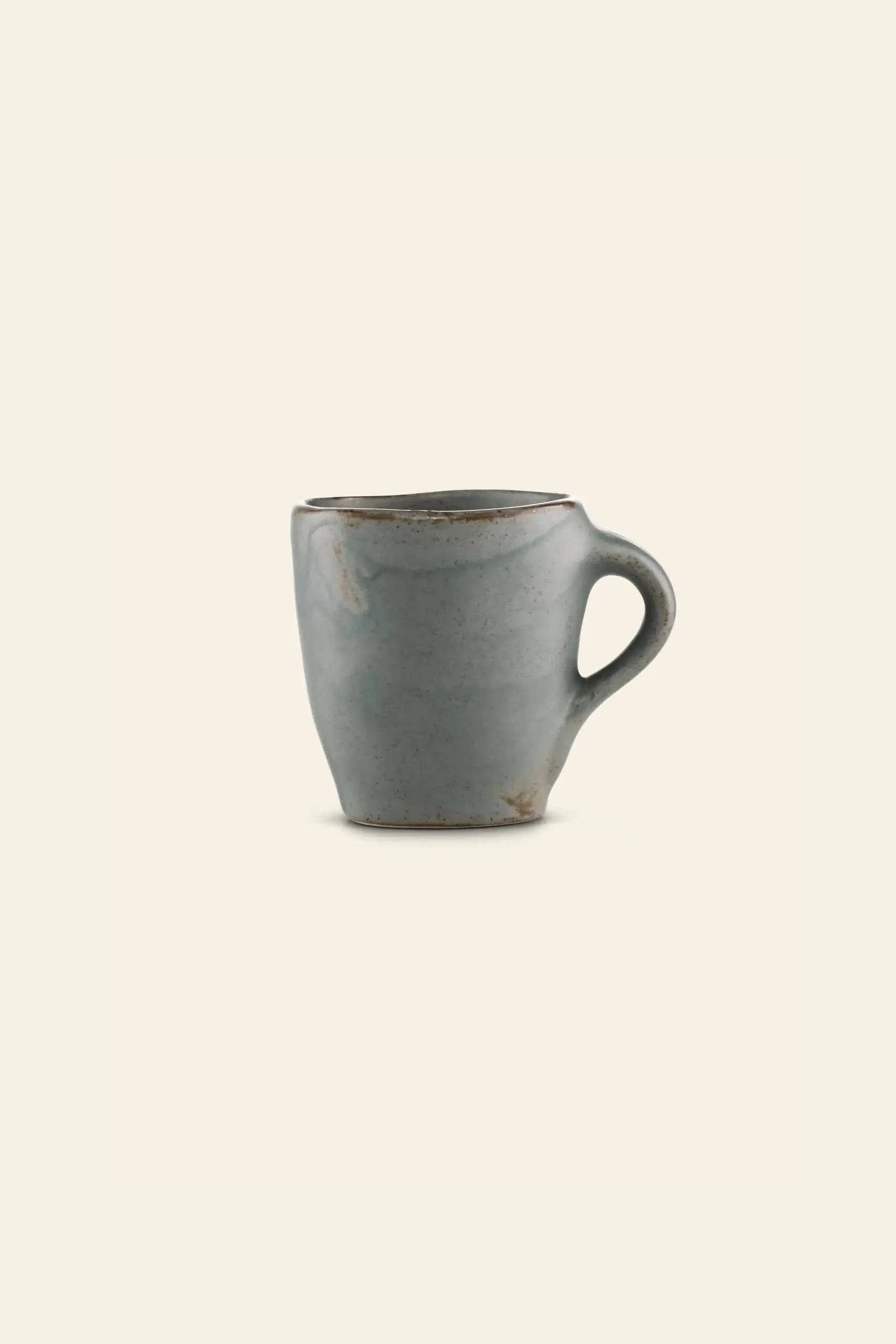 The Kra Sanctuary Karst Free-Form Coffee Mug by Kra Sanctuary features a small, handmade ceramic design with a curved handle, uneven rim, and muted gray glaze, exuding rustic charm on an off-white background.