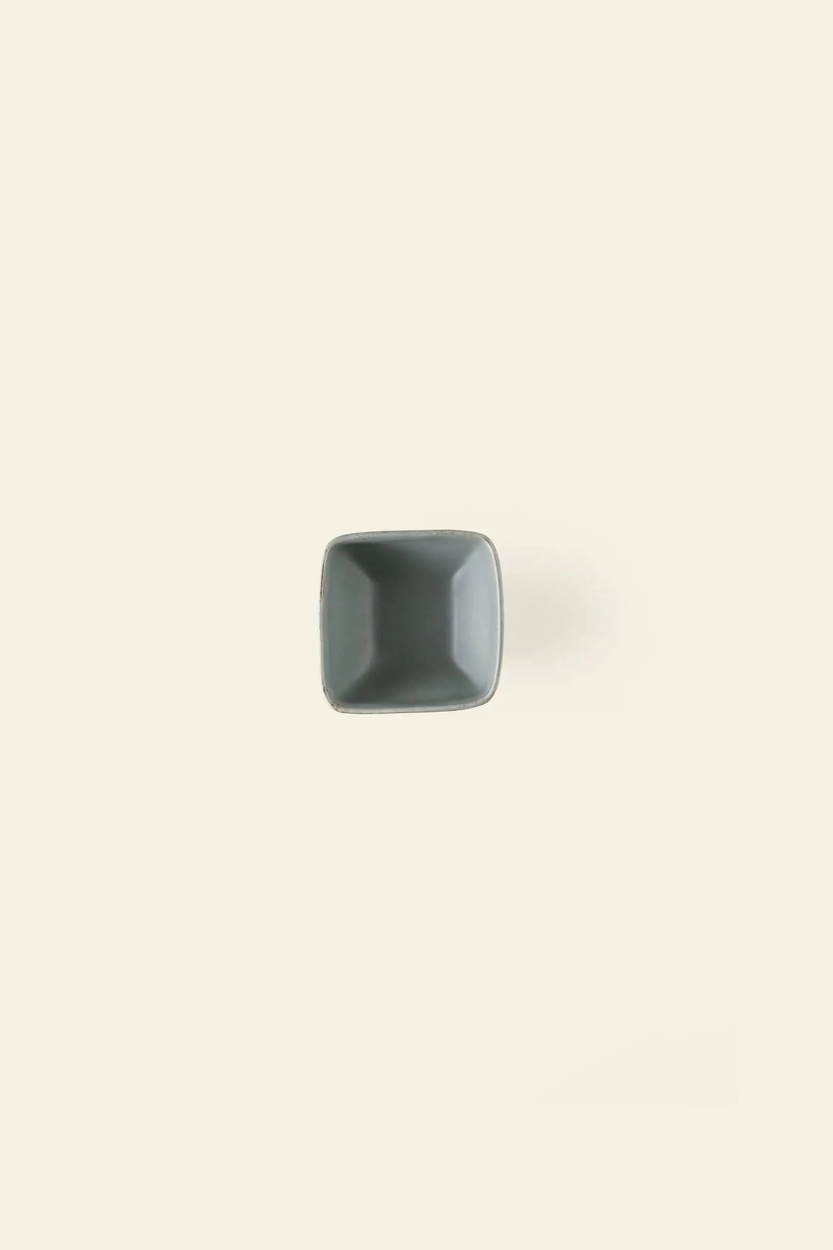 The Kra Sanctuary Karst Square Snack Bowl, a small empty gray bowl, is centered on a light beige background, casting a faint shadow to the right and highlighting its distinctive design.