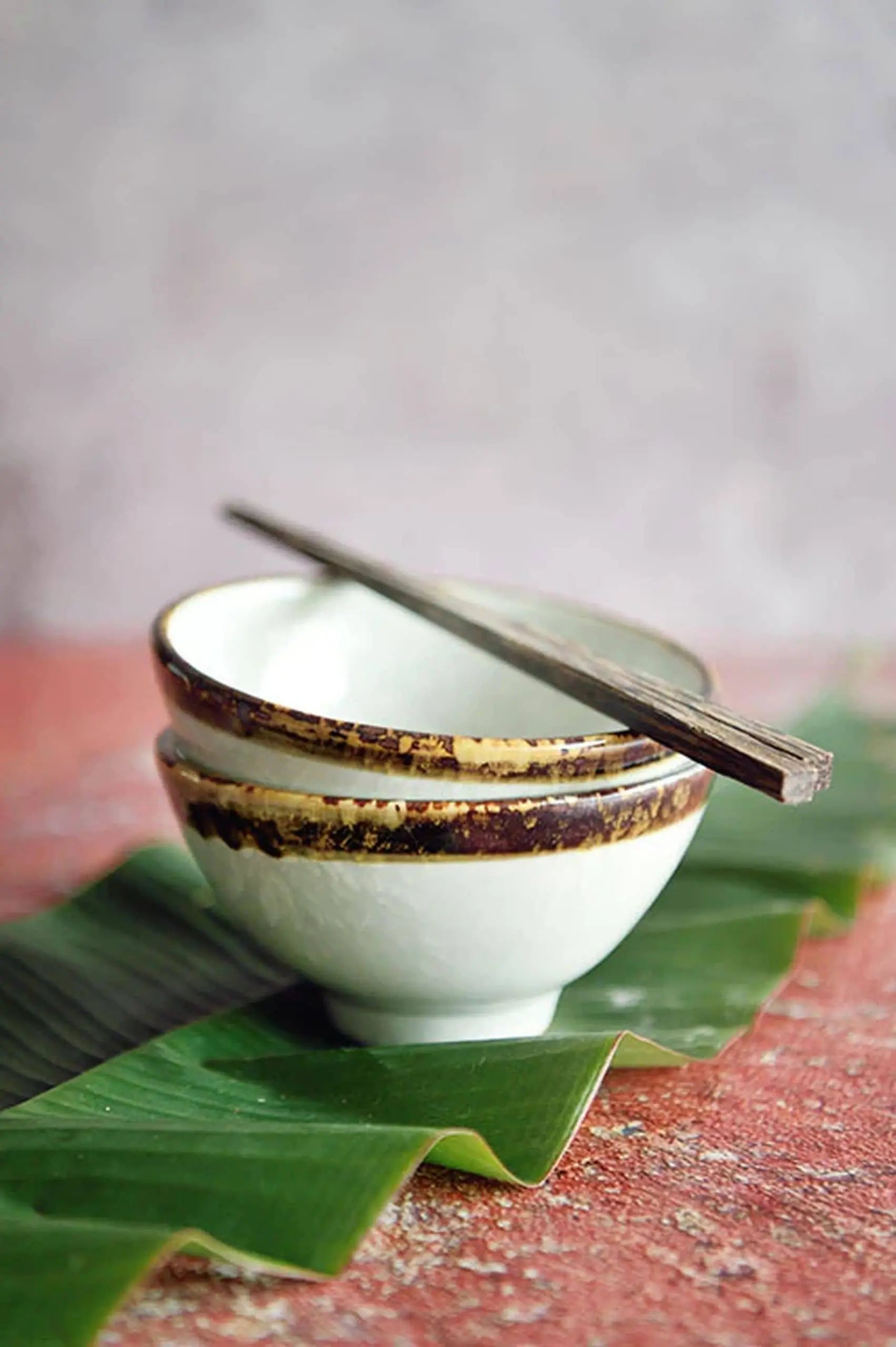 Two stacked Kra Sanctuary Satay Everyday Rice Bowls in white ceramic with brown rims rest on a green leaf, topped with wooden chopsticks. The blurred red and gray background enhances the inviting vibe. Brand: Kra Sanctuary.