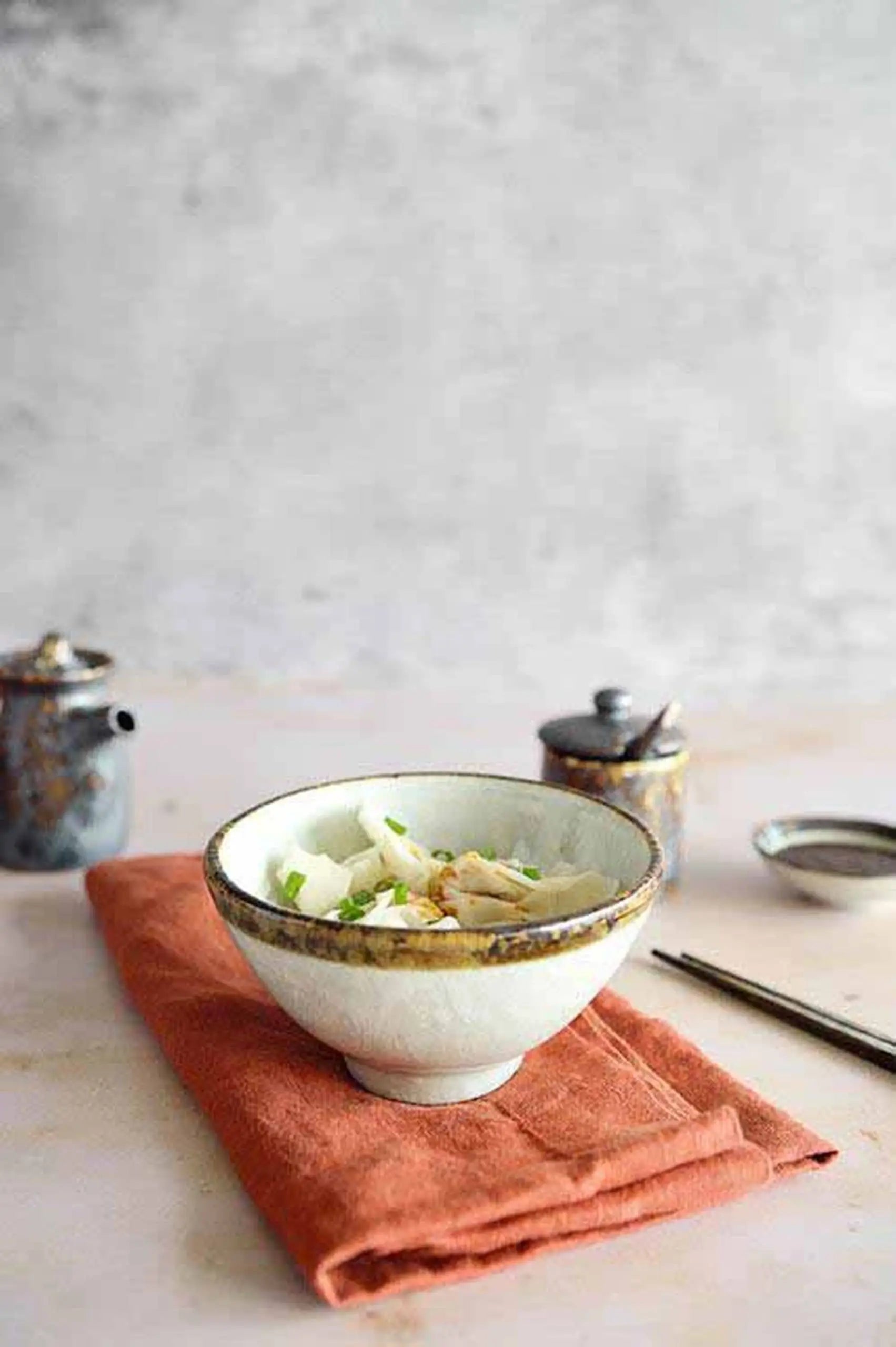 The Kra Sanctuary Satay Everyday Rice Bowl by Kra Sanctuary, filled with food and garnished with green onions, rests on an orange cloth with chopsticks and small jars nearby, enhancing your dining experience on a neutral backdrop.
