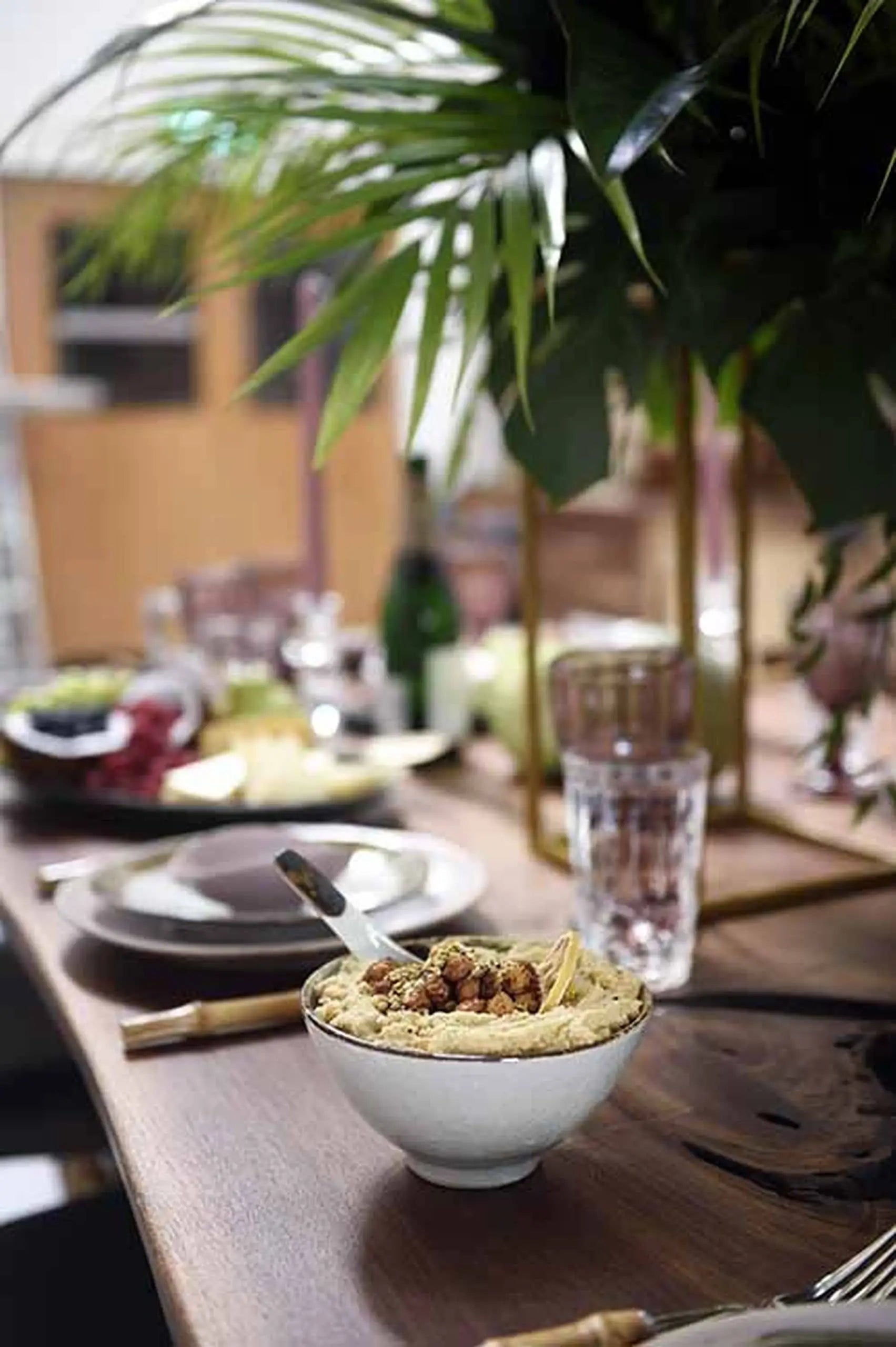 A close-up of a wooden dining table set with Kra Sanctuary Satay Everyday Rice Bowl dinnerware, plates, glasses, cutlery, and a white bowl of hummus. Green leafy plants add freshness for an inviting dining experience.