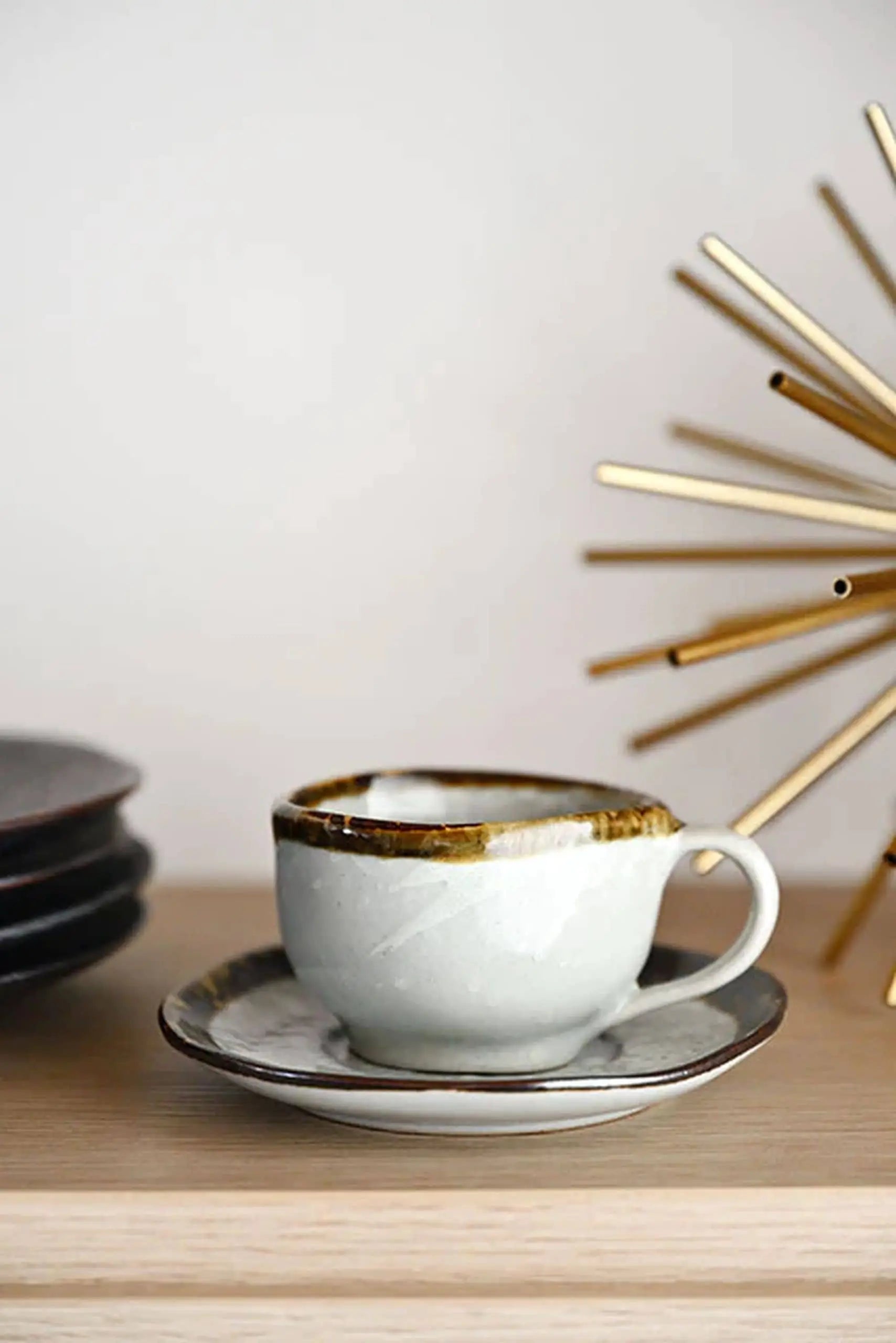 The Kra Sanctuary Satay Free-Form Cup and Coaster from Kra Sanctuary features a white ceramic cup with an iridescent border on a matching coaster, displayed beside earthy plates and a gold starburst sculpture on a wooden surface.