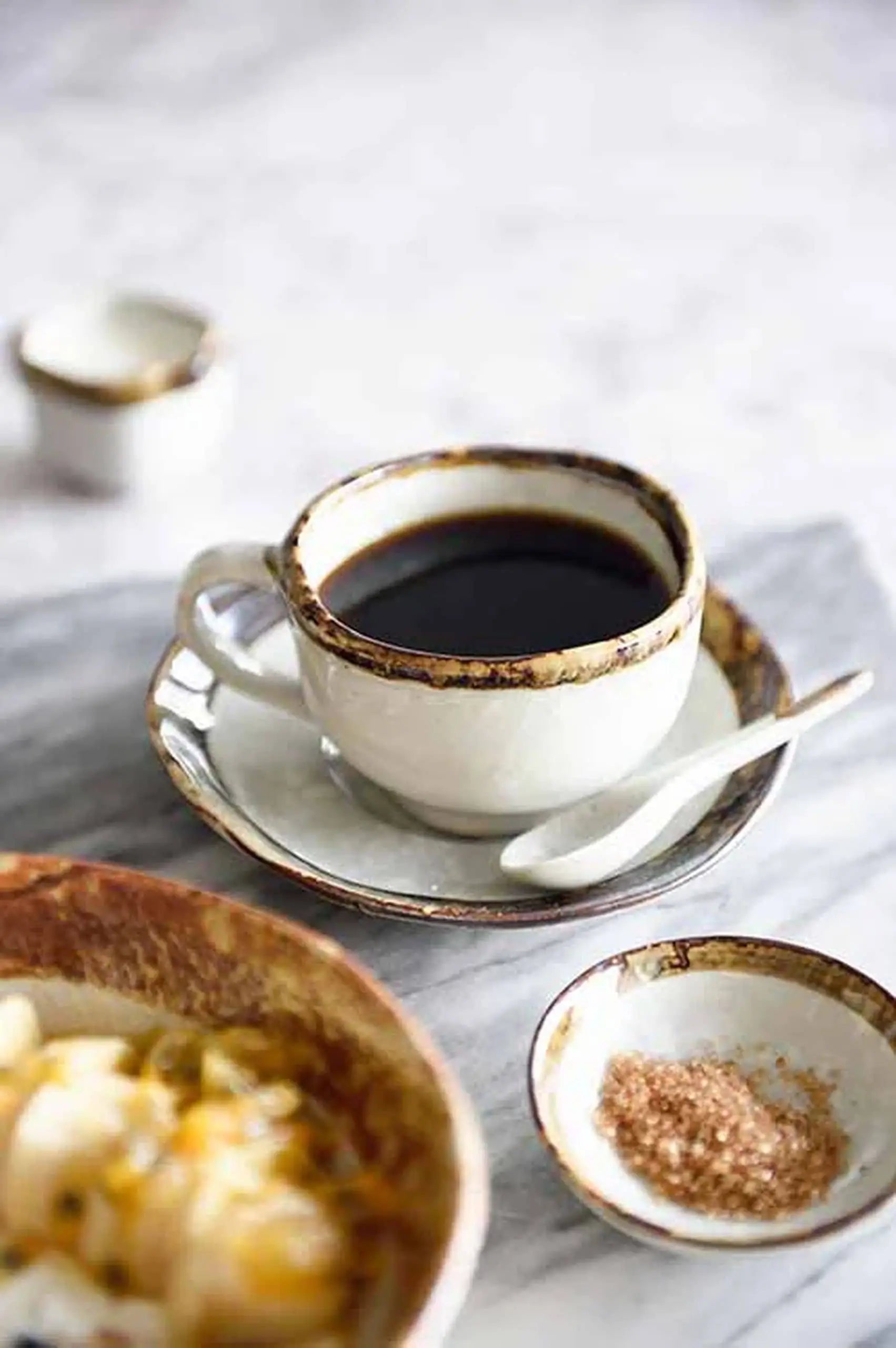 The Kra Sanctuary Satay Free-Form Cup and Coaster by Kra Sanctuary holds black coffee, paired with a matching coaster and white spoon. A brown sugar dish and part of a food bowl rest nearby, all displayed on a marble surface.
