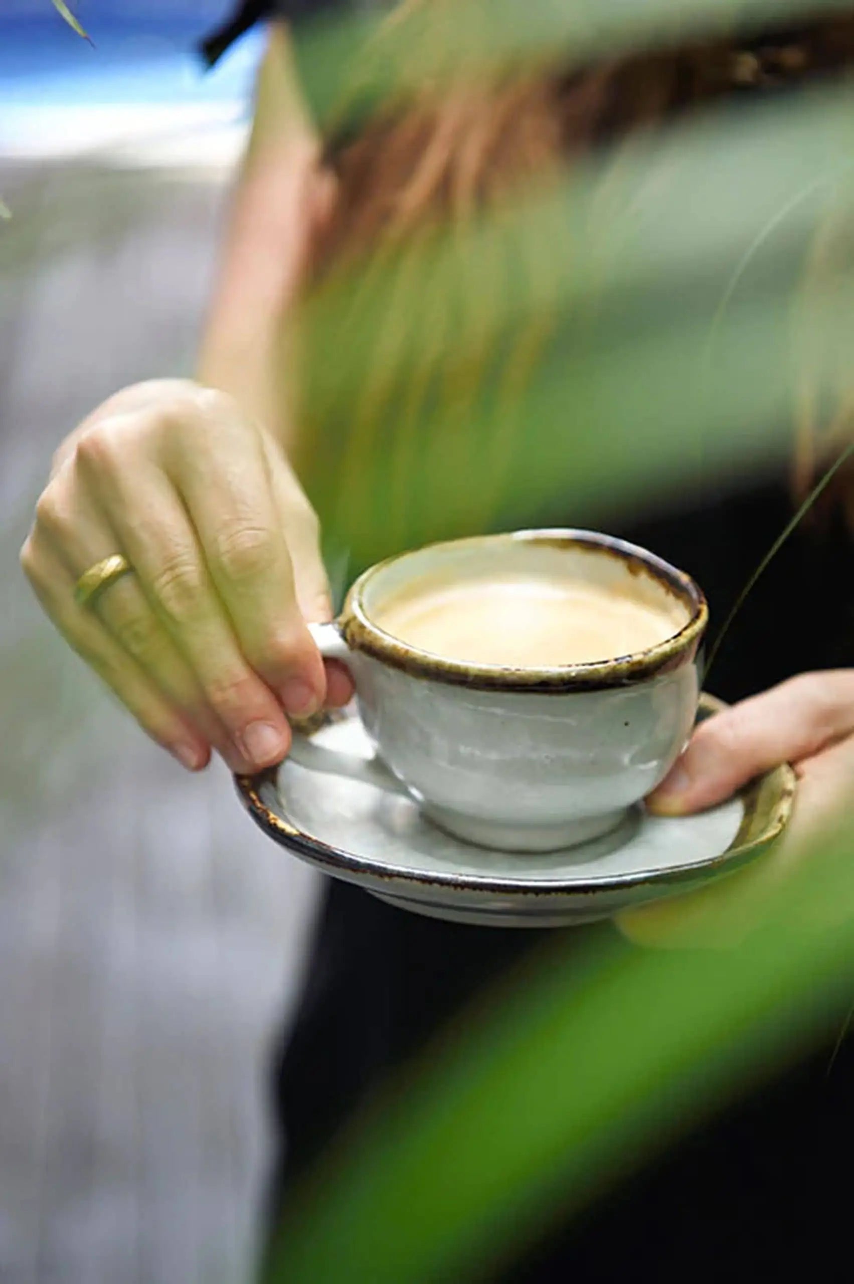 A person holds a Kra Sanctuary Satay Free-Form Cup with its matching coaster, both partially hidden by green leaves. They wear a gold ring, and the softly blurred background adds an earthy ambiance to the Kra Sanctuary setting.